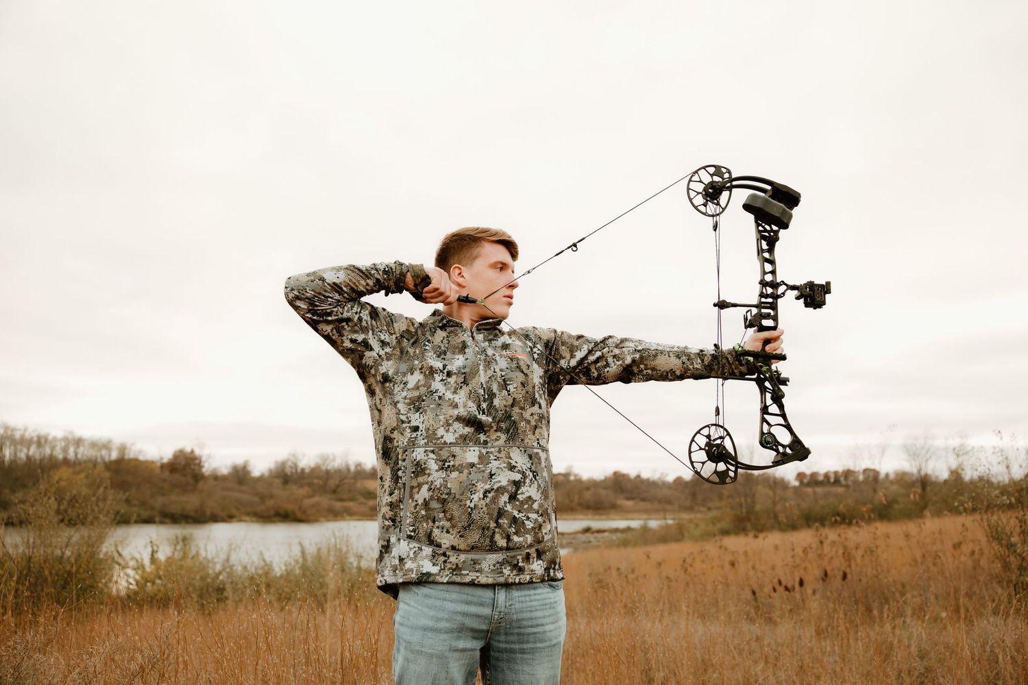 High school senior in camouflage clothing aims compound bow in open field during sunset.