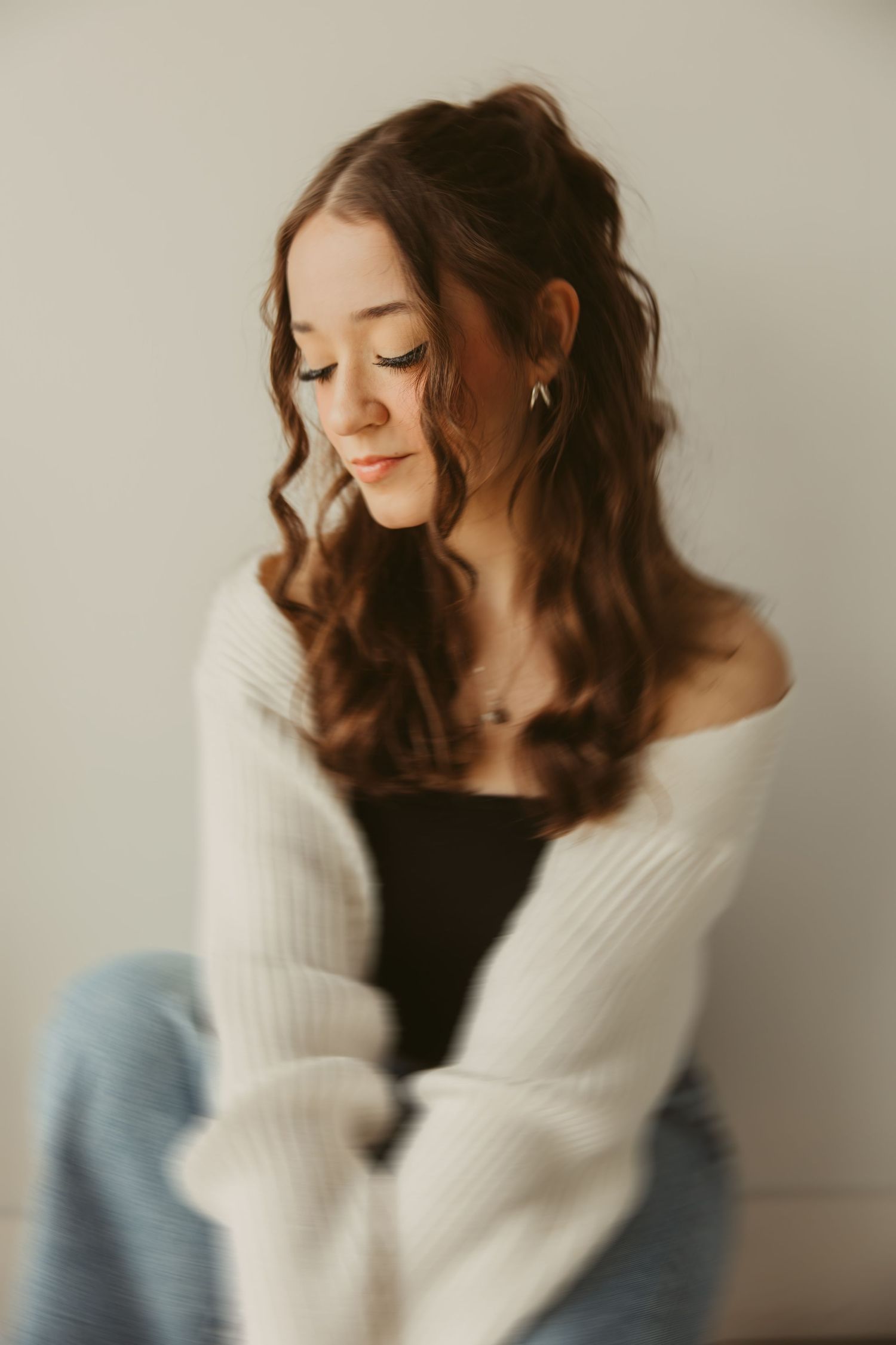 Side profile portrait of a high school senior with long wavy brown hair wearing a white sweater and jeans against neutral background.