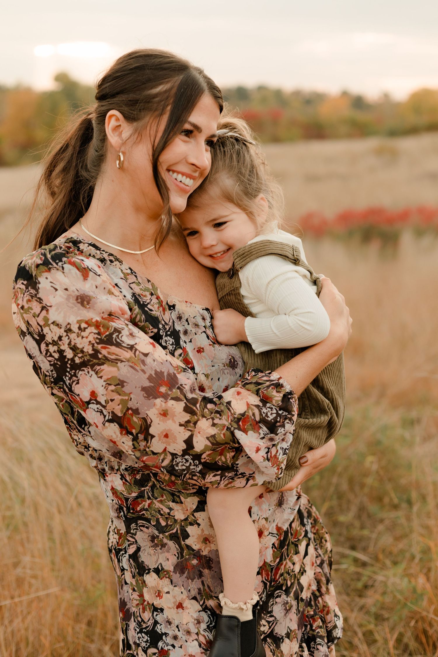 A mother and daughter share a tender hug and smile together in an autumn field at sunset while wearing floral dress and overall dress.
