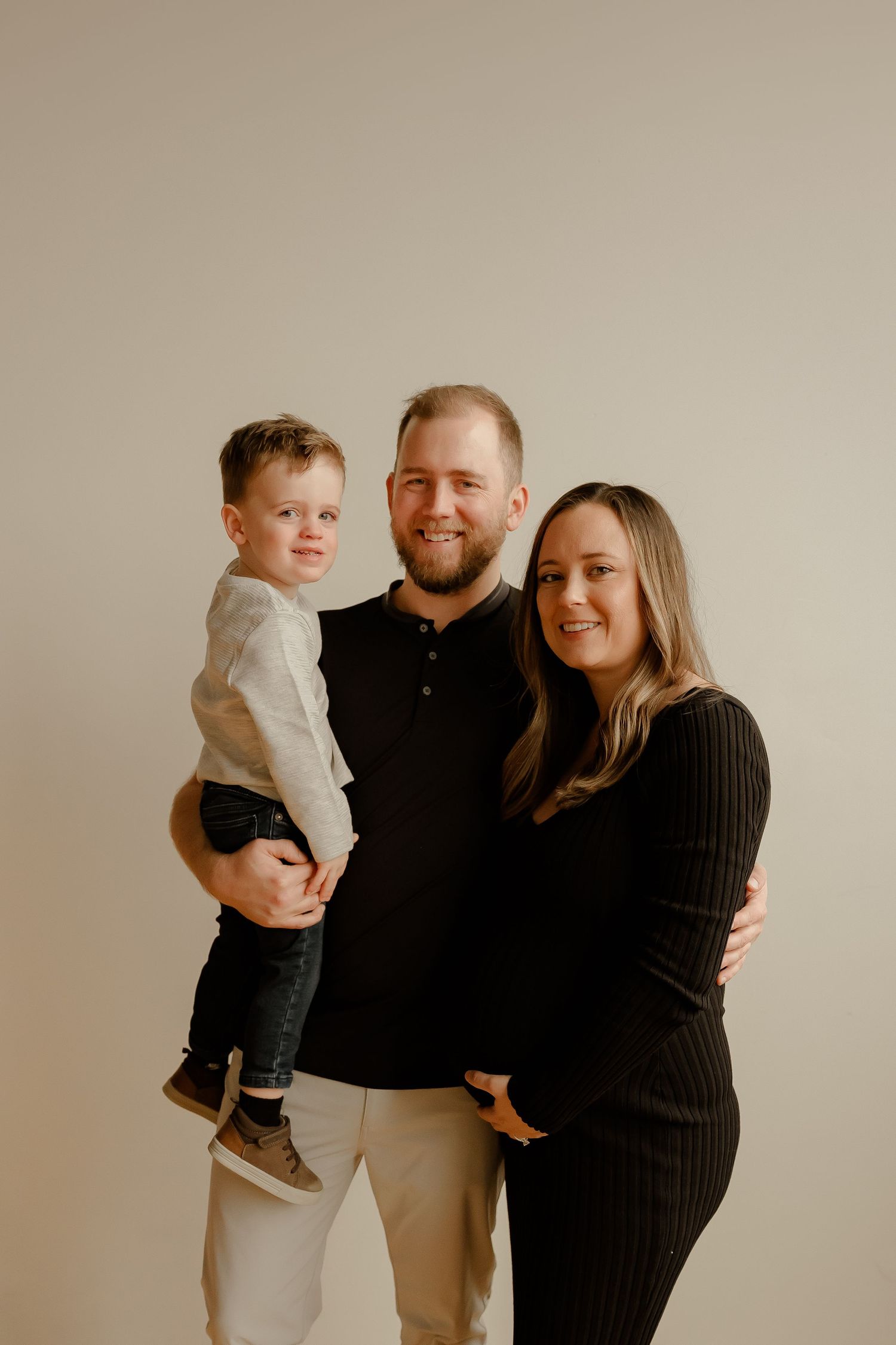 A family poses together for a portrait against a neutral beige background wearing coordinated black and neutral clothing.