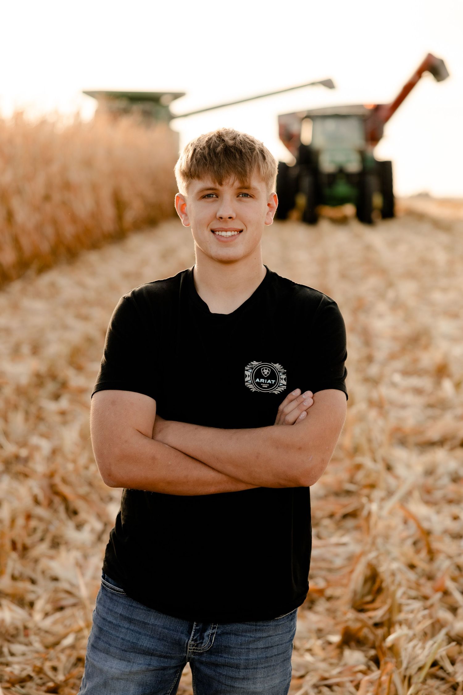 High school senior in black t-shirt stands in harvested field with combine and tractor working in background at sunset.