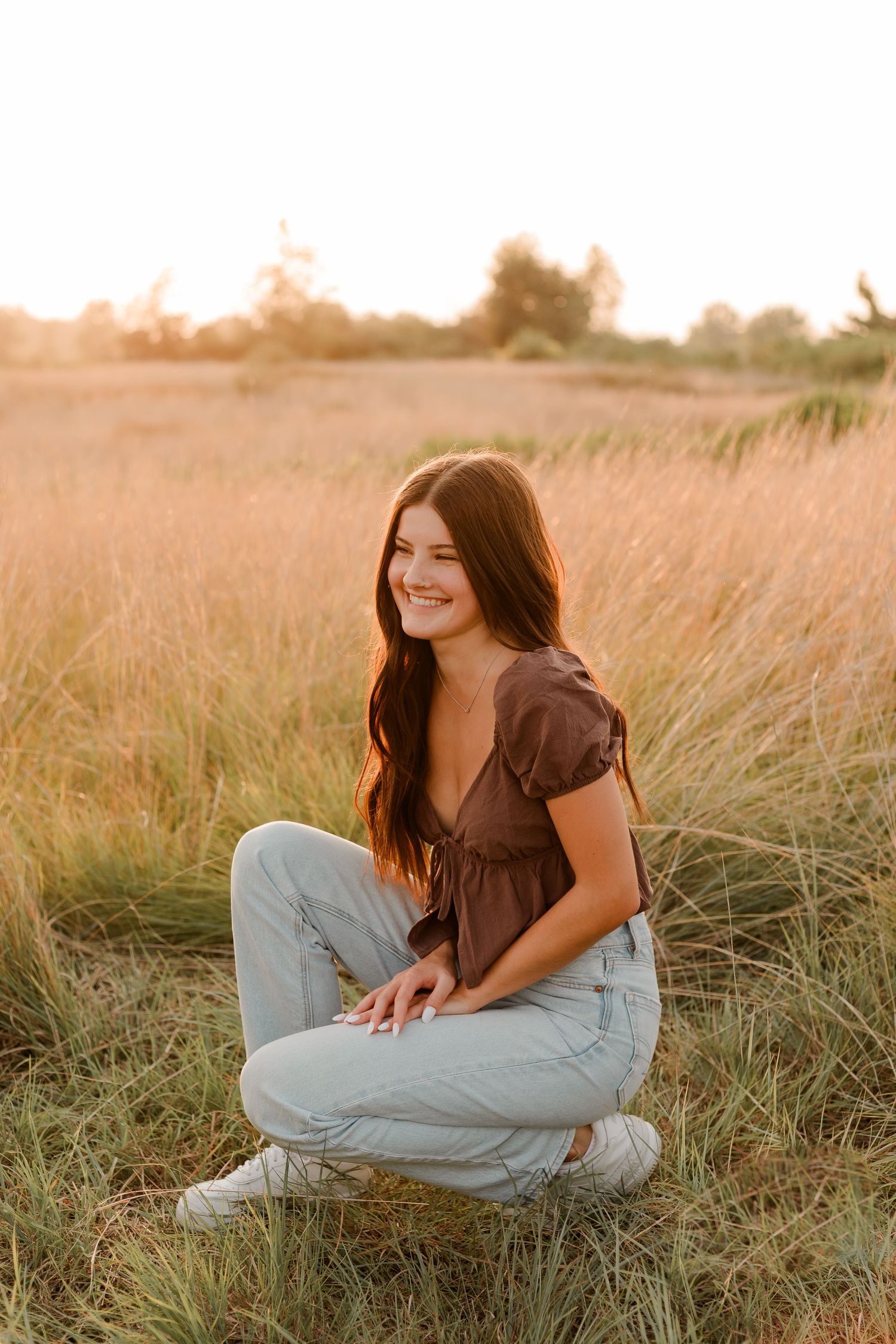 A high school senior in a brown top and light jeans smiles while sitting in a golden field at sunset.
