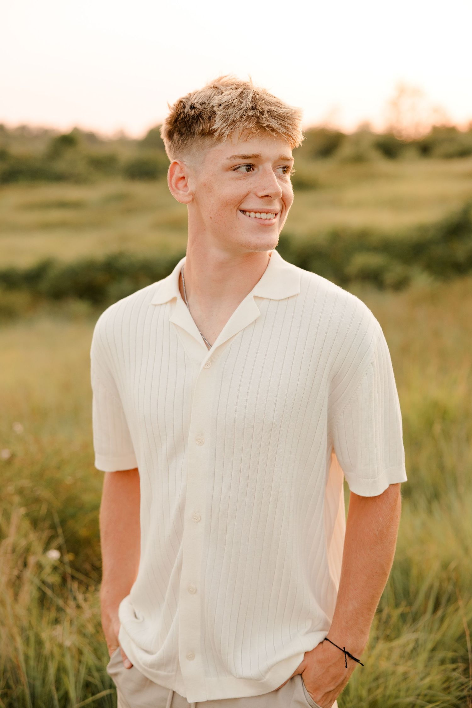 A high school senior smiling in a white collared shirt stands in a golden field during sunset.
