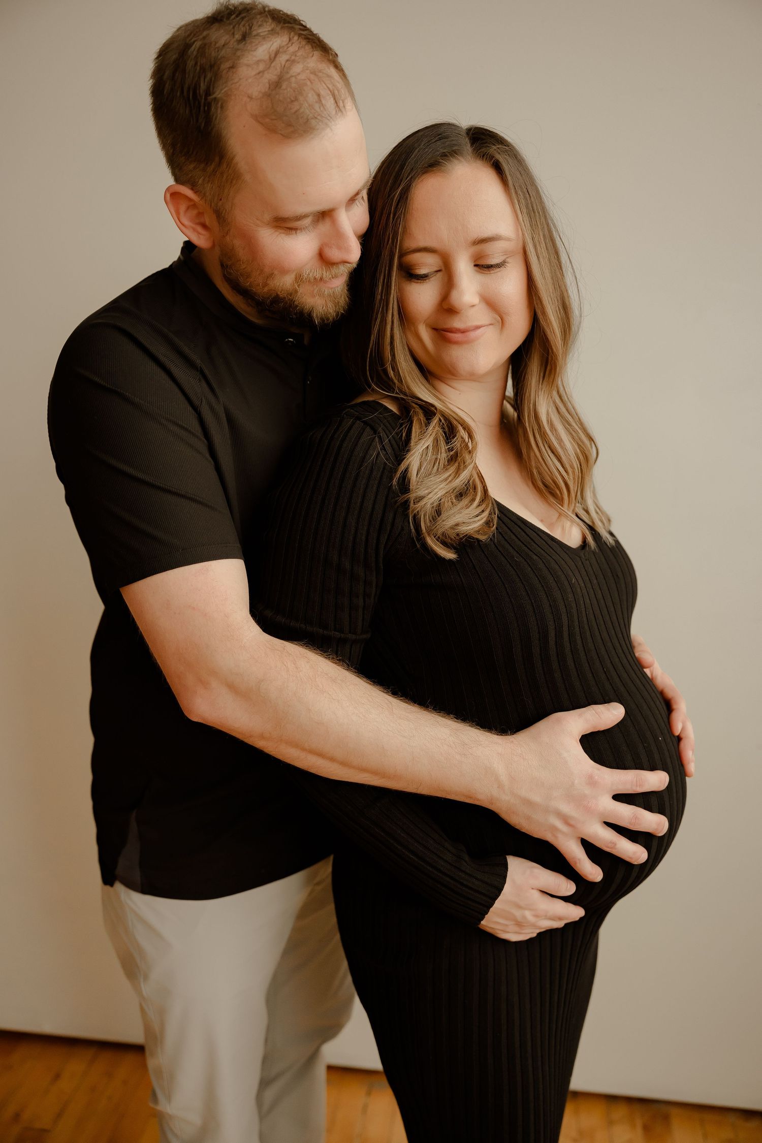 Intimate maternity photo showing a couple in black embracing with hands on the pregnant belly against beige background.