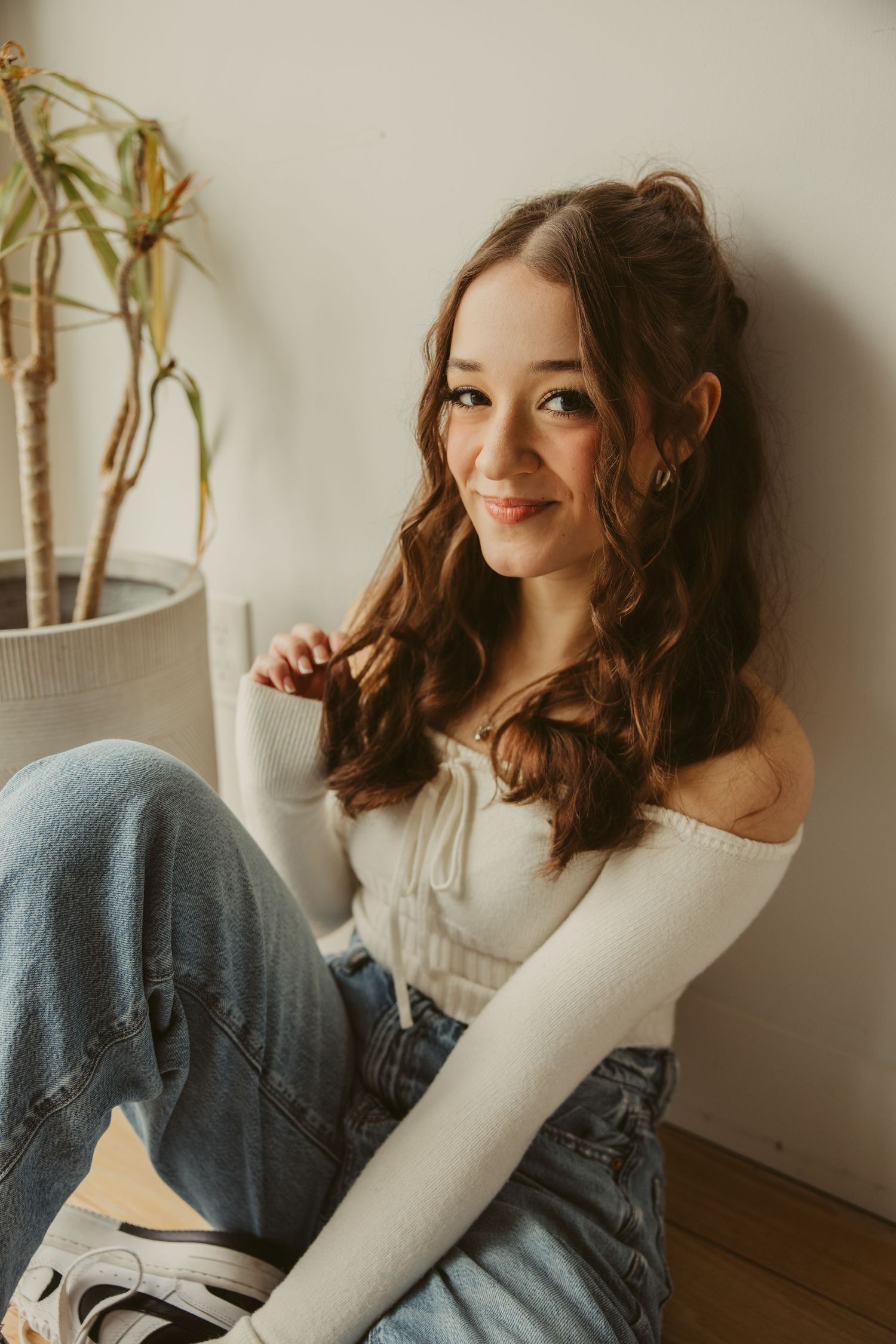 High school senior in white off-shoulder top and jeans sits casually by potted bamboo plant in natural lighting.
