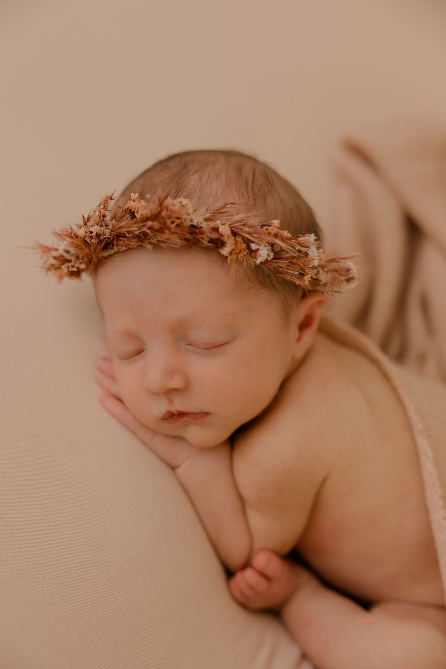 Peaceful sleeping newborn wearing delicate autumn-colored dried flower crown against beige backdrop.