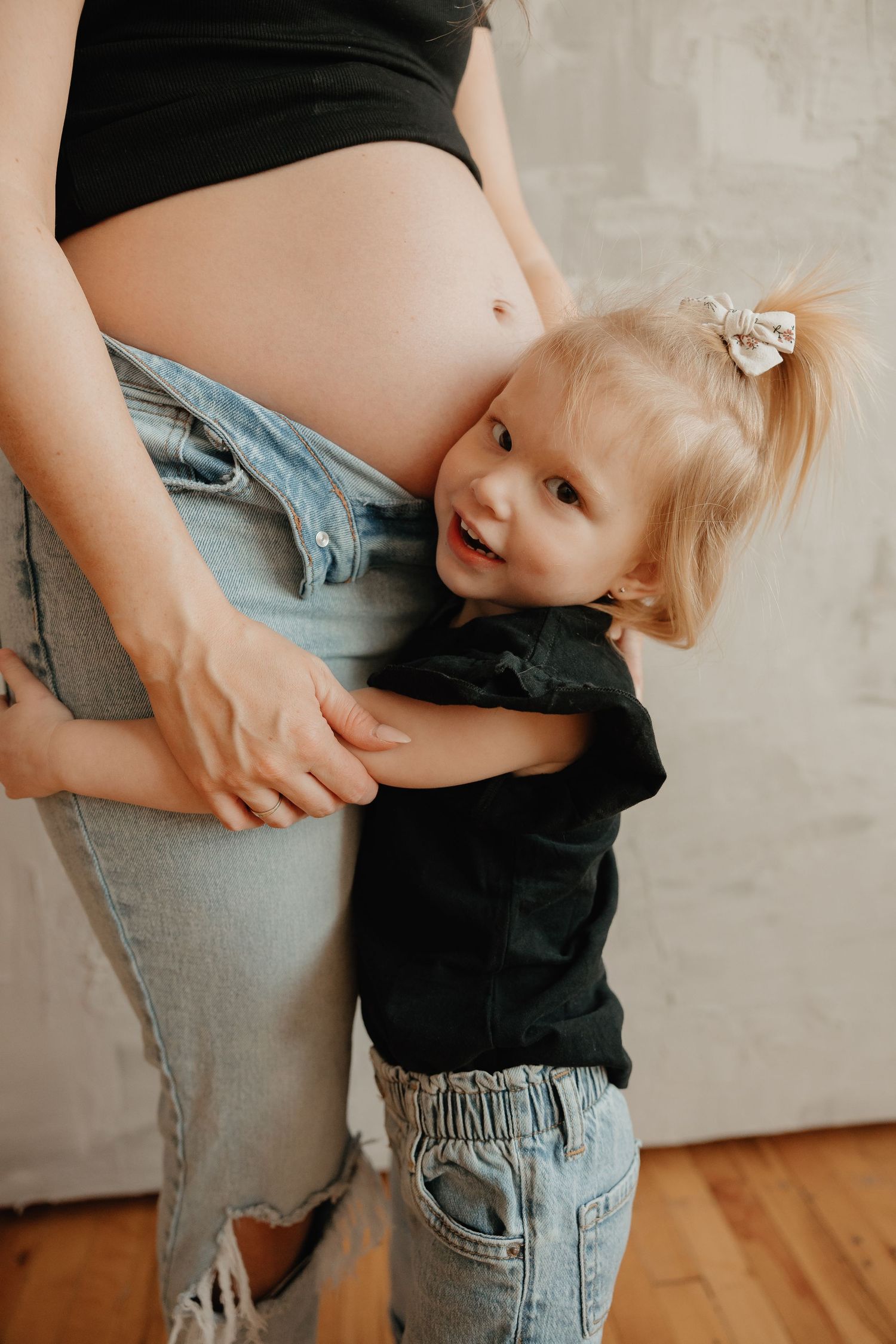 Toddler in denim hugging pregnant mother's belly while wearing black shirt against gray wall background.