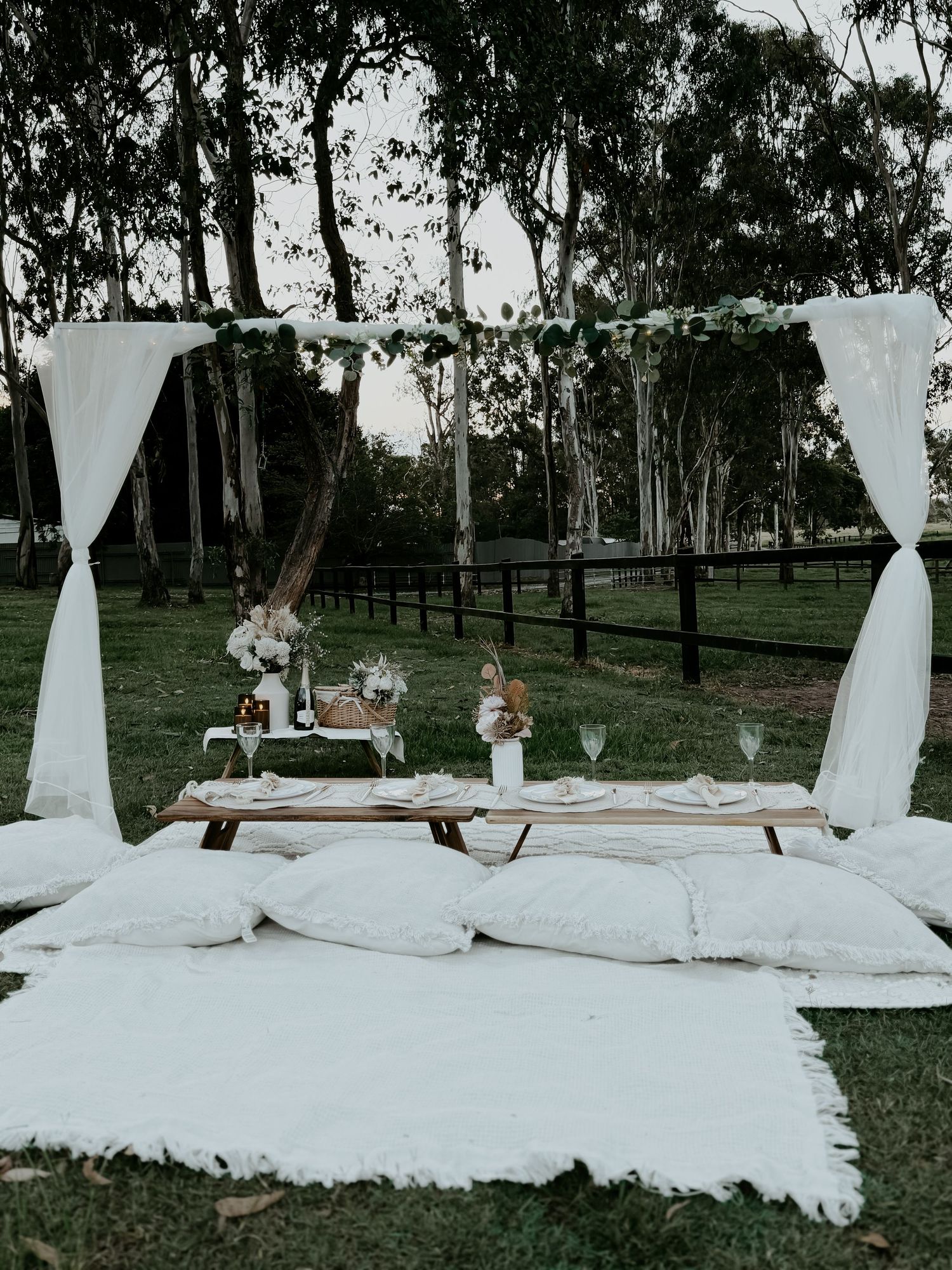 Rustic outdoor picnic setup with white cushions, draping fabric, and wooden benches under trees at dusk.