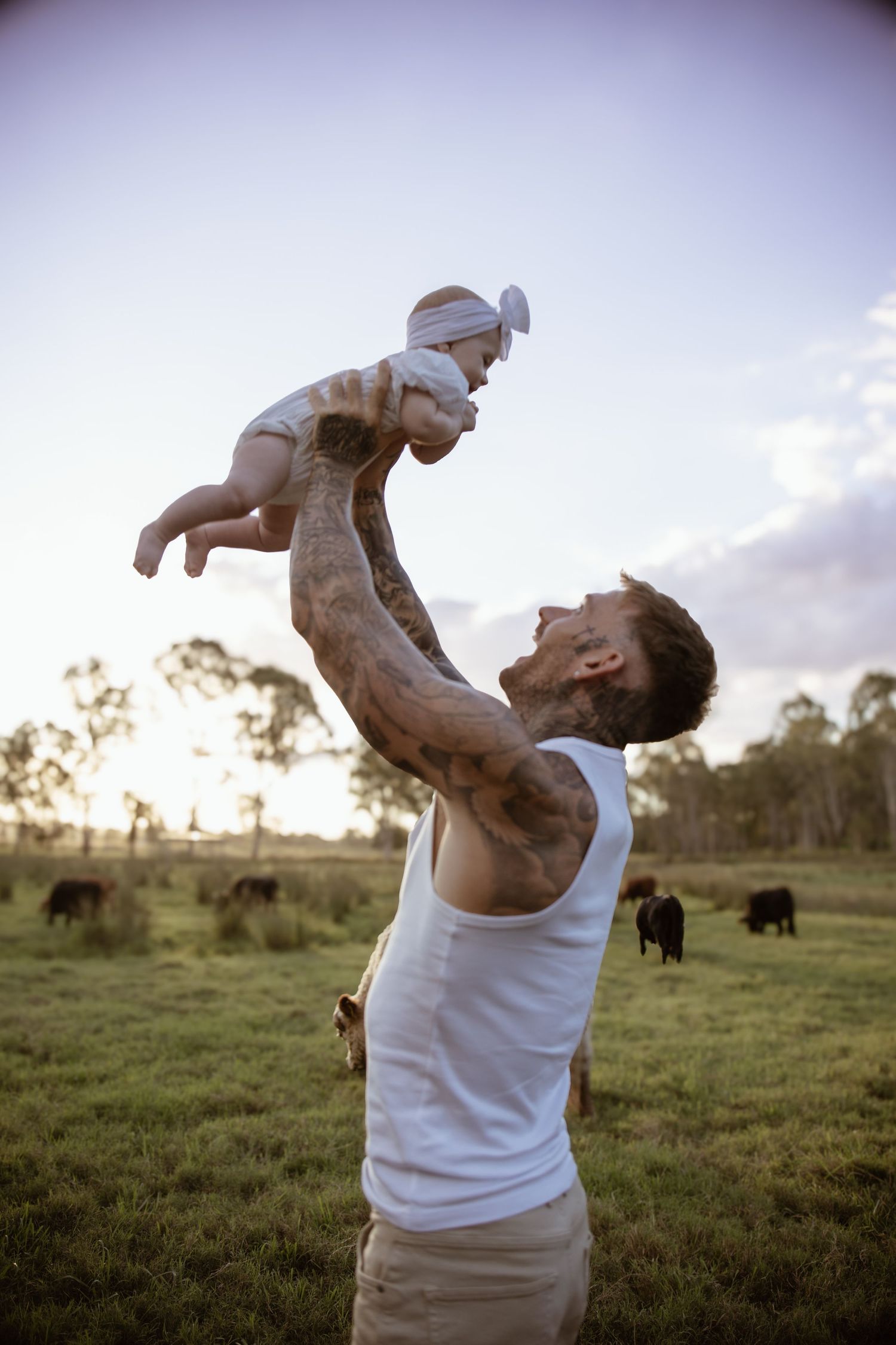 A parent in white tank top lifts a baby into the air at sunset in a field with cattle grazing in the background.