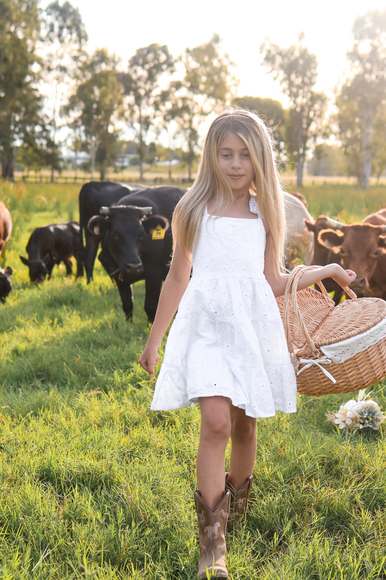 Girl walking through pasture in white dress with picnic basket and cows.