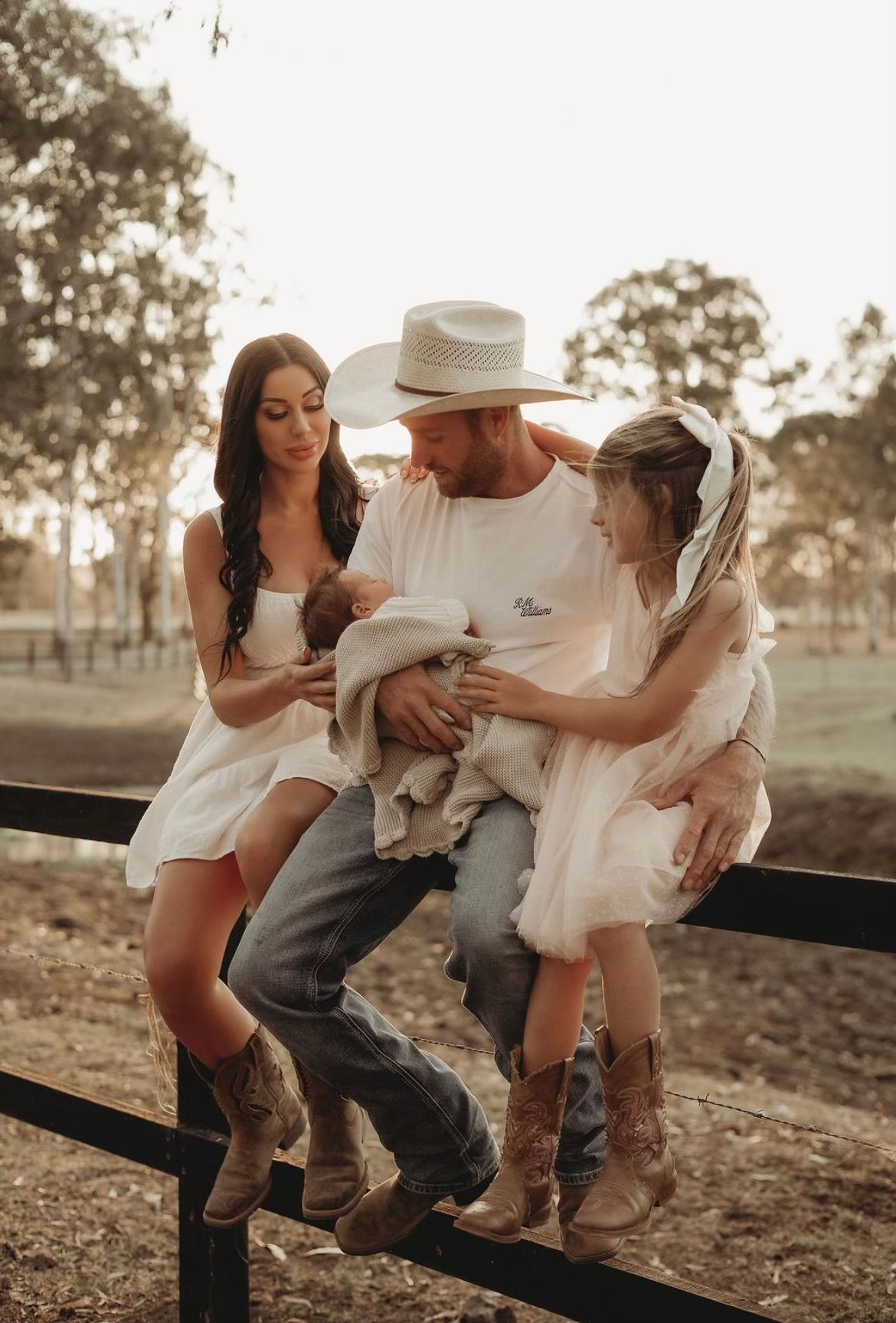 A family in white outfits and cowboy boots sits together on a rustic wooden fence at sunset in a country setting.