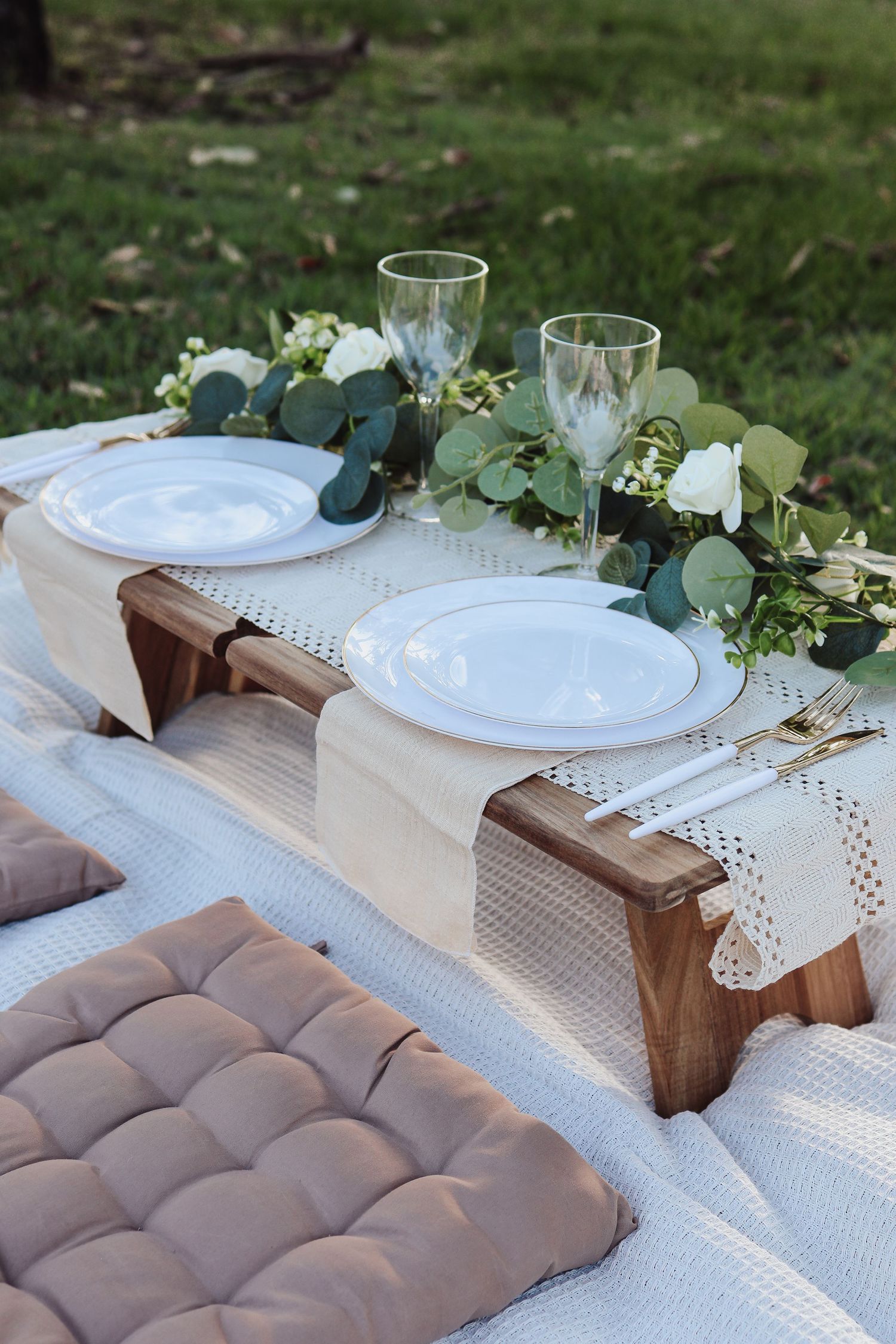 Elegant picnic table setting with white plates, wine glasses, and eucalyptus garland on a white tablecloth with brown cushions.