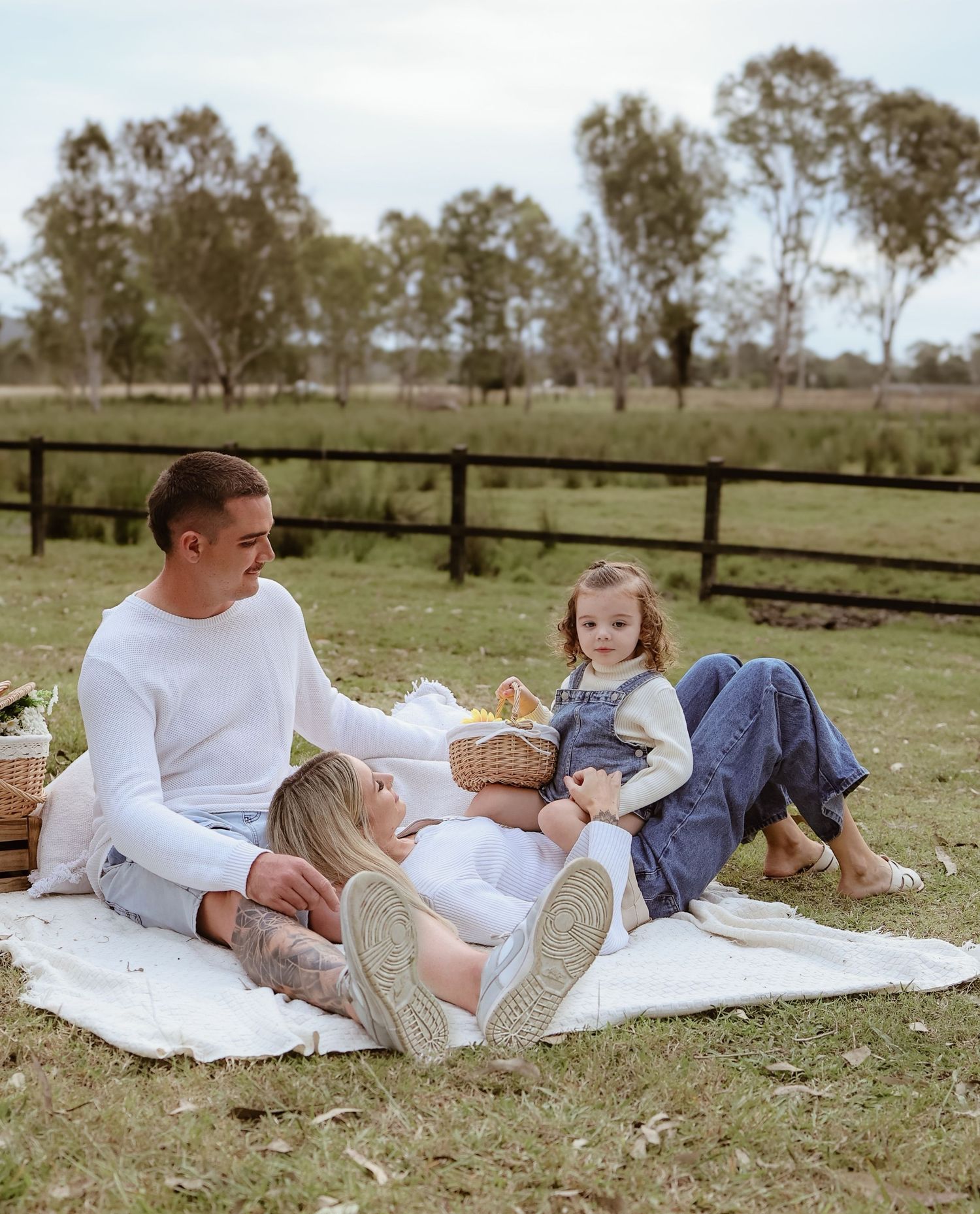 A family enjoys a relaxing picnic on a white blanket by a wooden fence in a rural countryside setting with eucalyptus trees.
