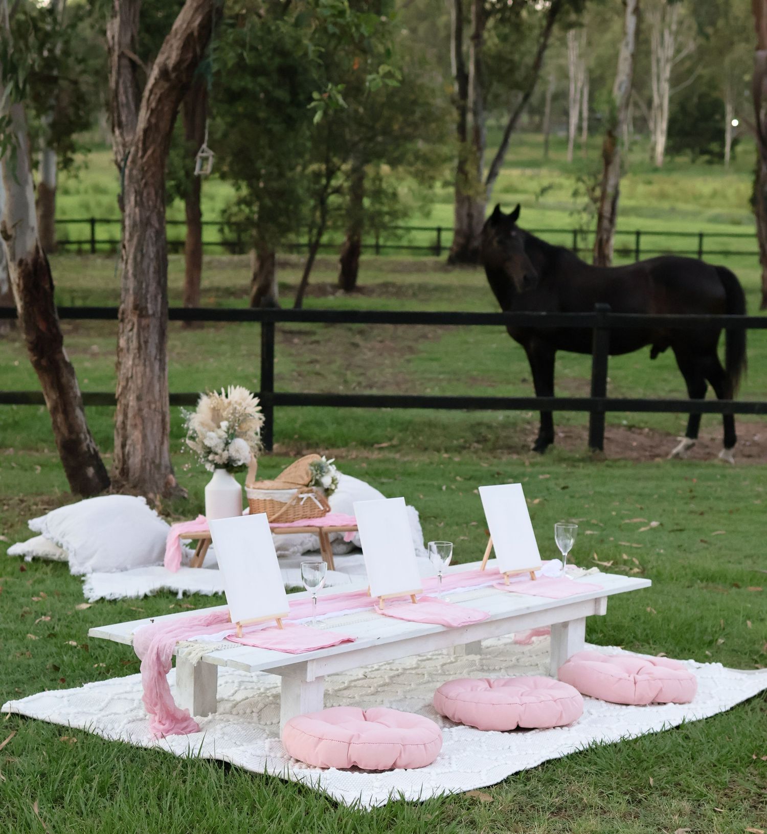 A romantic pink picnic setup with floor cushions and blankets in a green field with a black horse in the background.