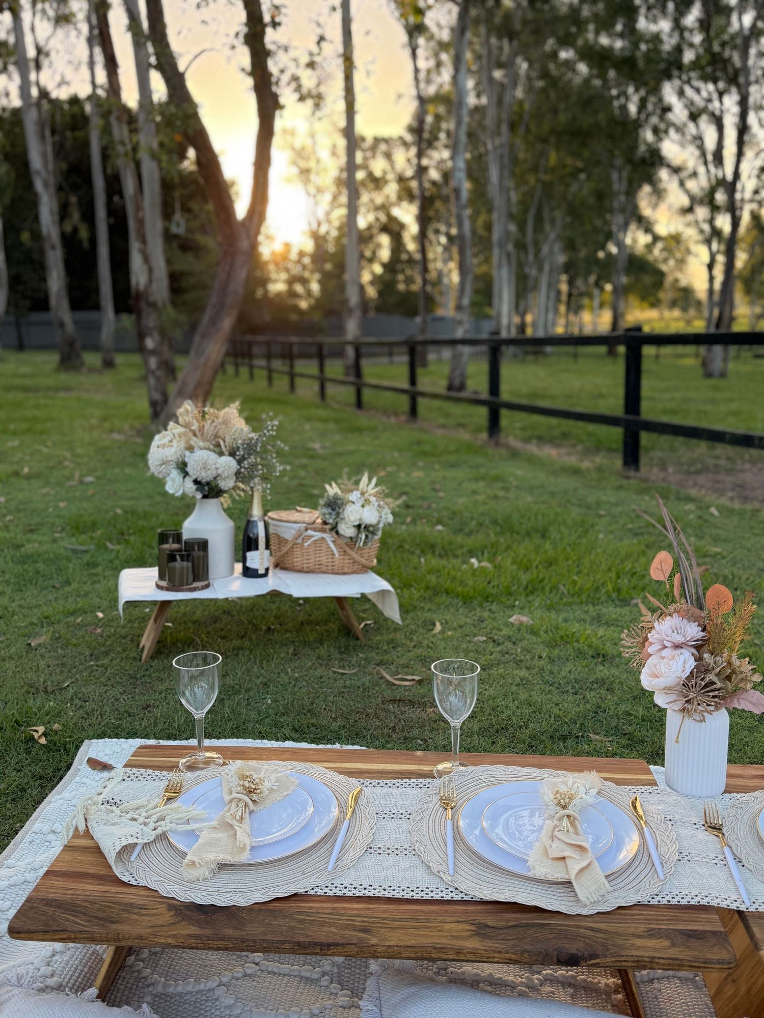 Rustic outdoor dining setup with wooden table, elegant place settings and floral arrangements at sunset behind birch trees.