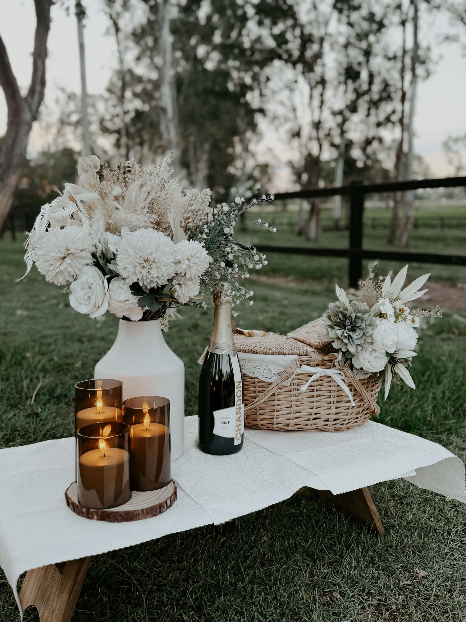 A rustic outdoor picnic setup with white flowers in a vase, lit candles, champagne bottle and wicker basket on grass.