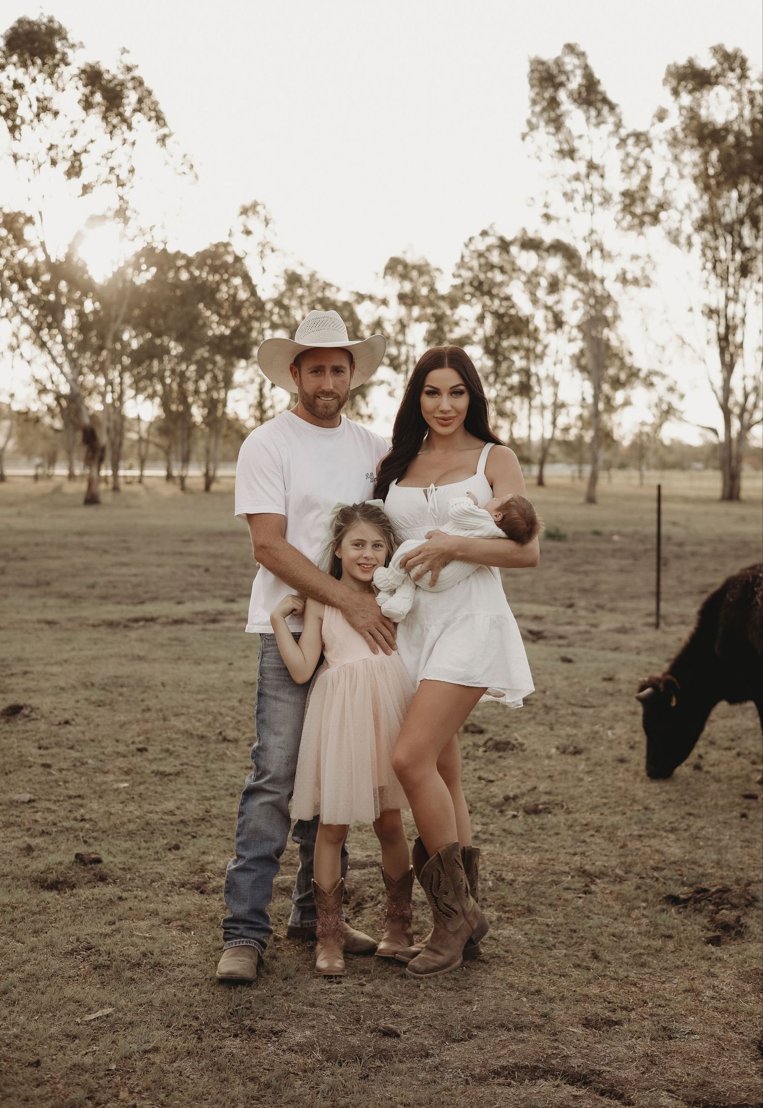 A family poses together in a rural setting at sunset with eucalyptus trees in the background and a cow visible to the side.