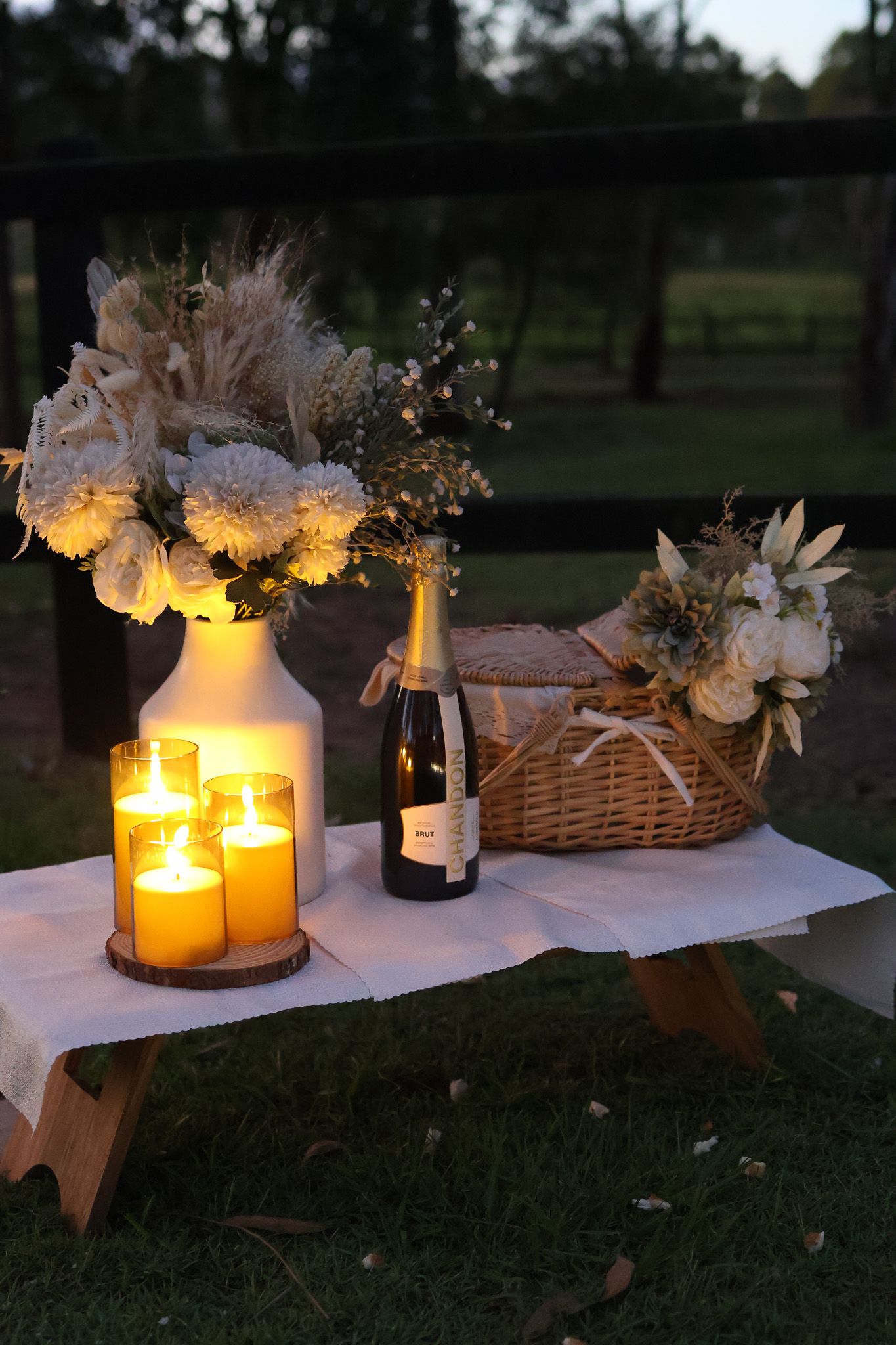 Evening outdoor display with illuminated flowers, candles, and picnic basket on wooden platform.