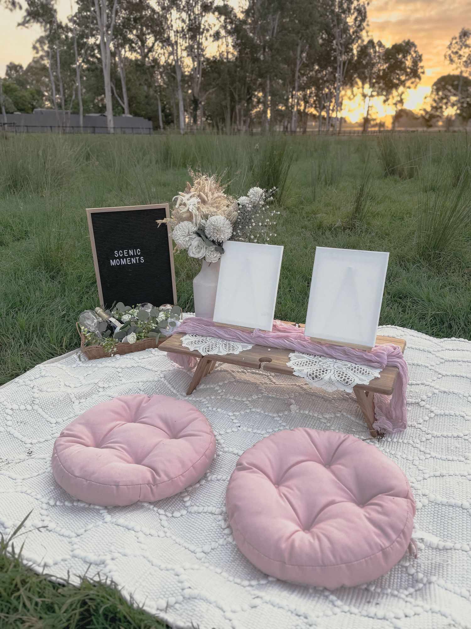 Outdoor picnic setup with pink round cushions, white tablecloth, and decorative display stands against sunset and trees.