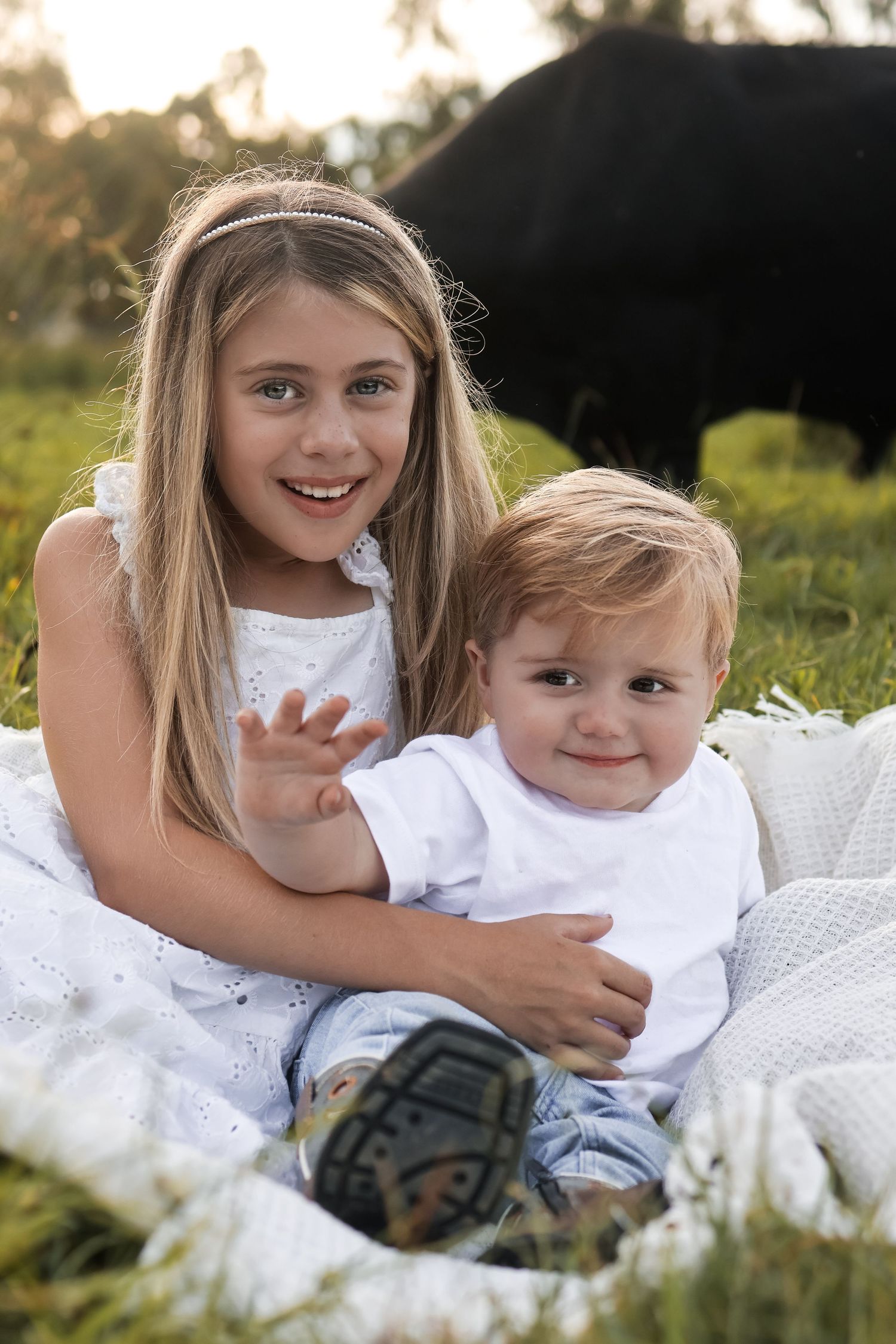 Sweet moment of siblings sitting together on white blanket with cows in background.