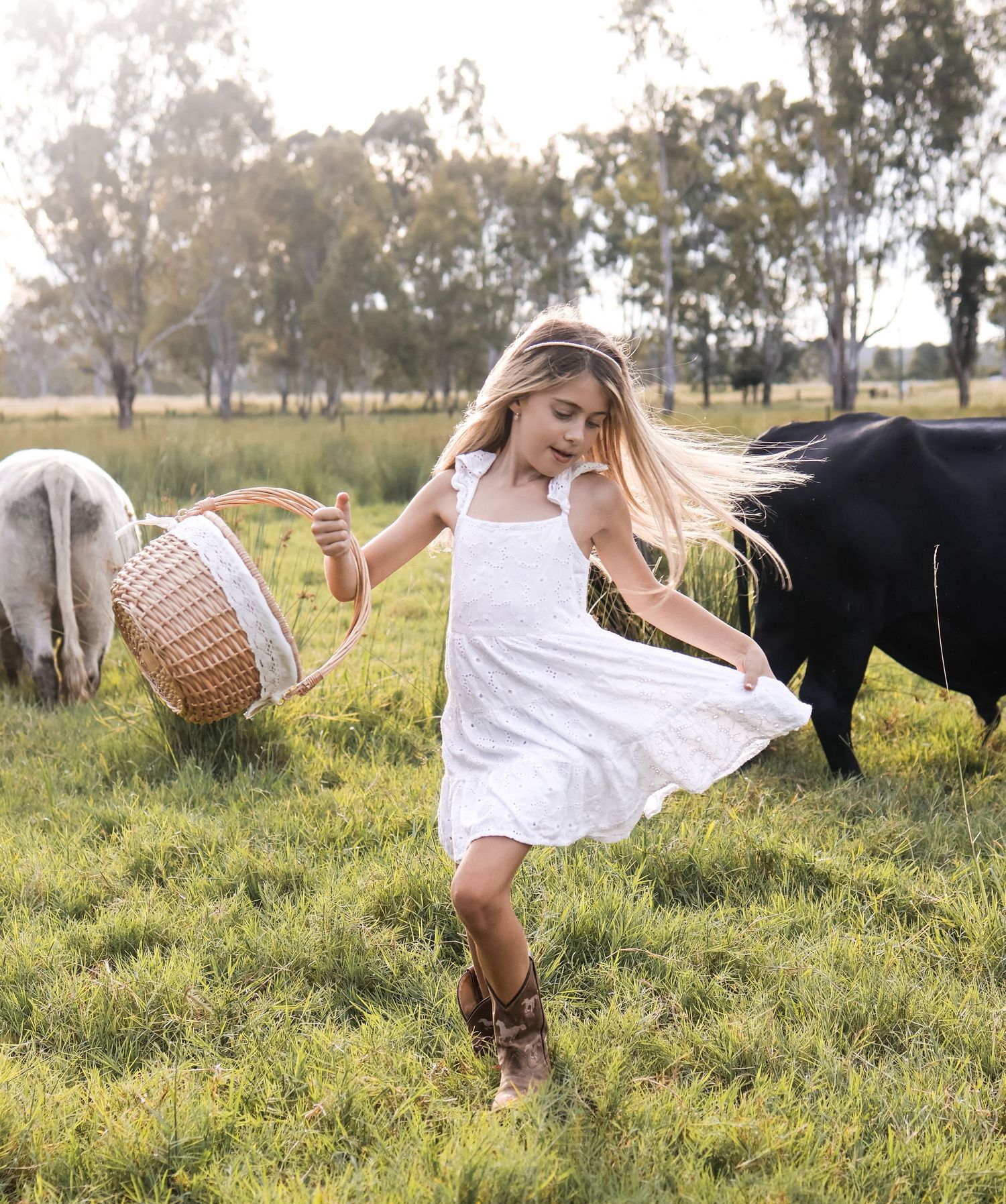 Girl in white dress twirling in countryside field with picnic basket and farm animals.