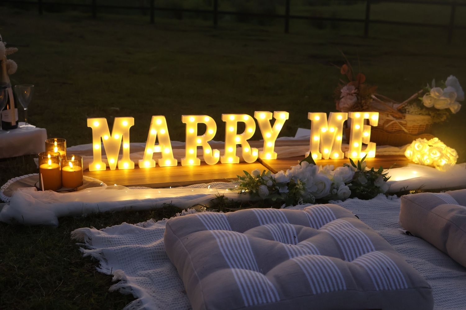 Illuminated 'MARRY ME' letters glow against a dark background with cozy floor cushions and romantic candle lighting.