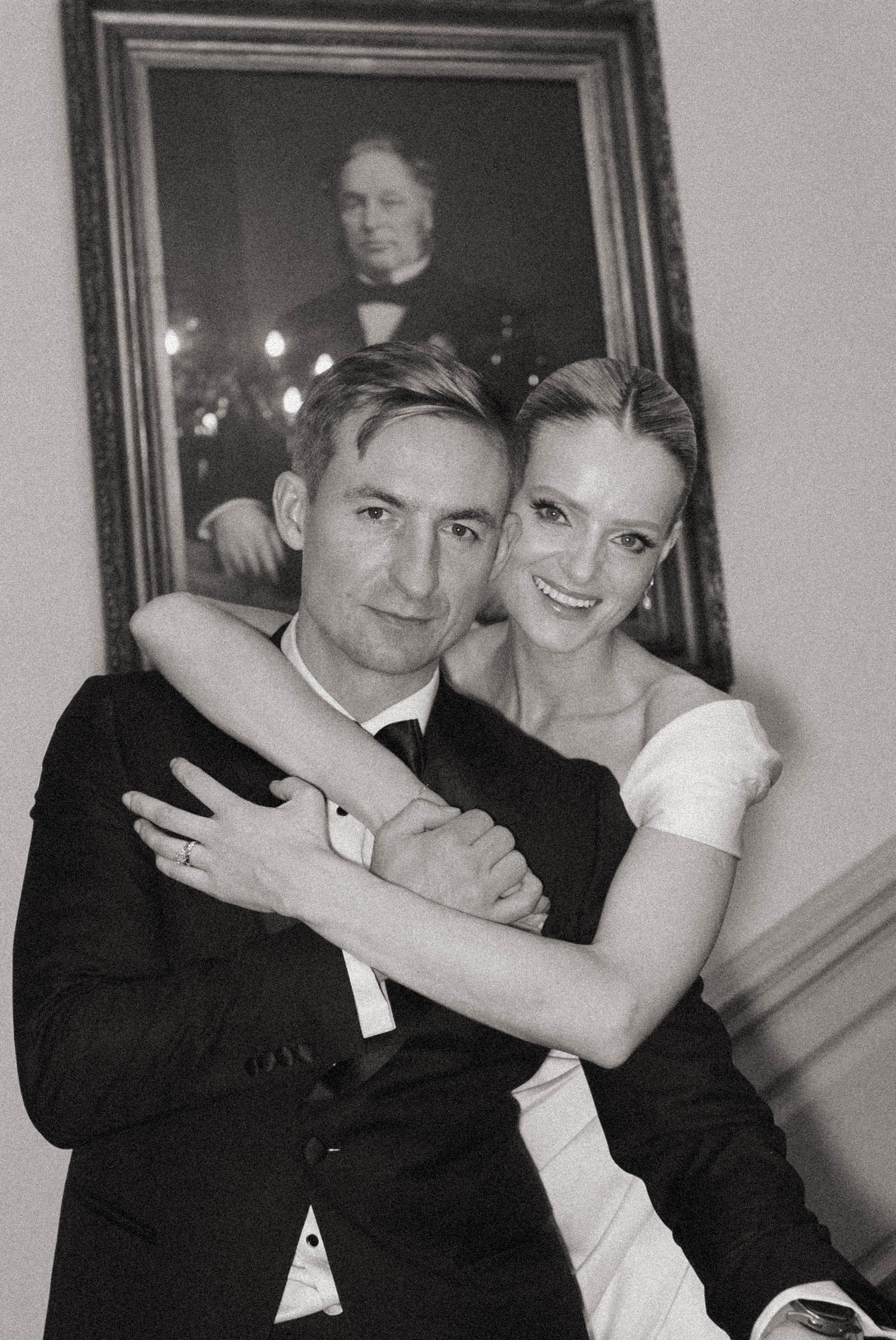 A black and white wedding photo shows a couple in formal attire embracing with a portrait visible in the background. happy smiles weddings T