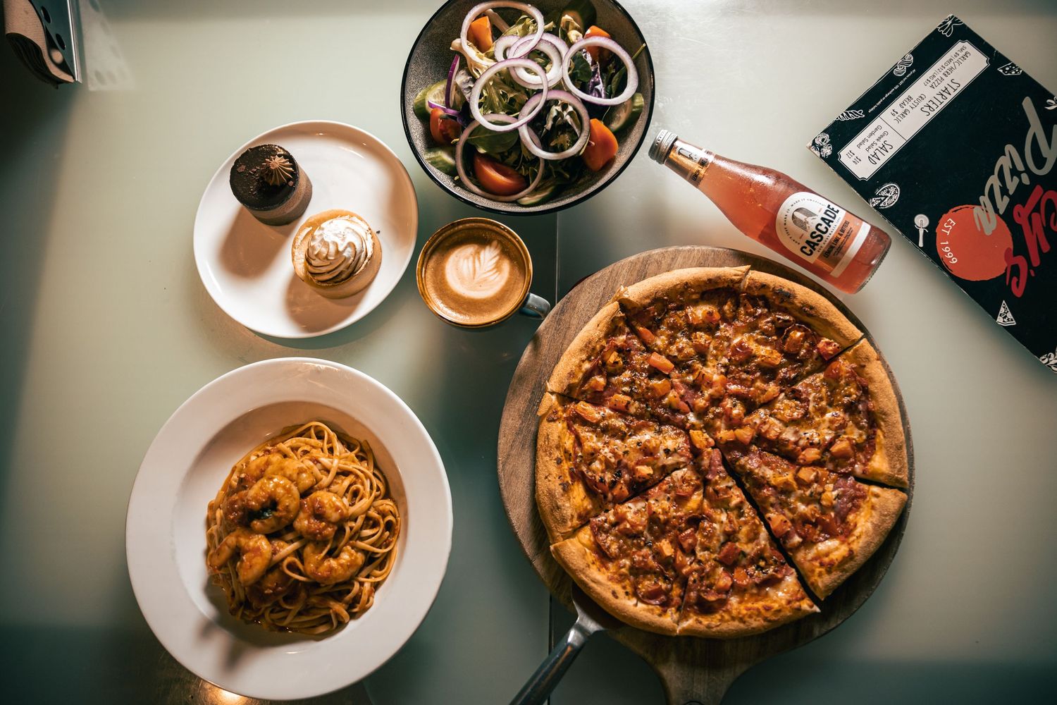 Overhead flat lay of pizza, pasta, drinks, and cutlery on dining table.