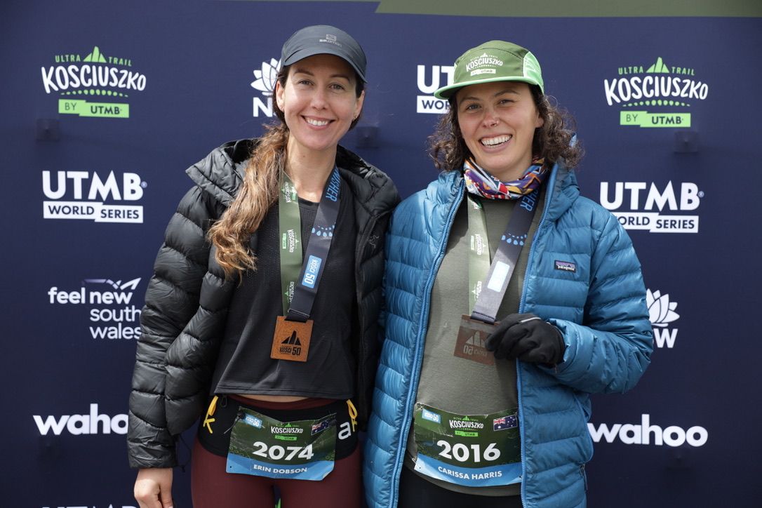 Two runners posing at event backdrop, smiling with race medals.