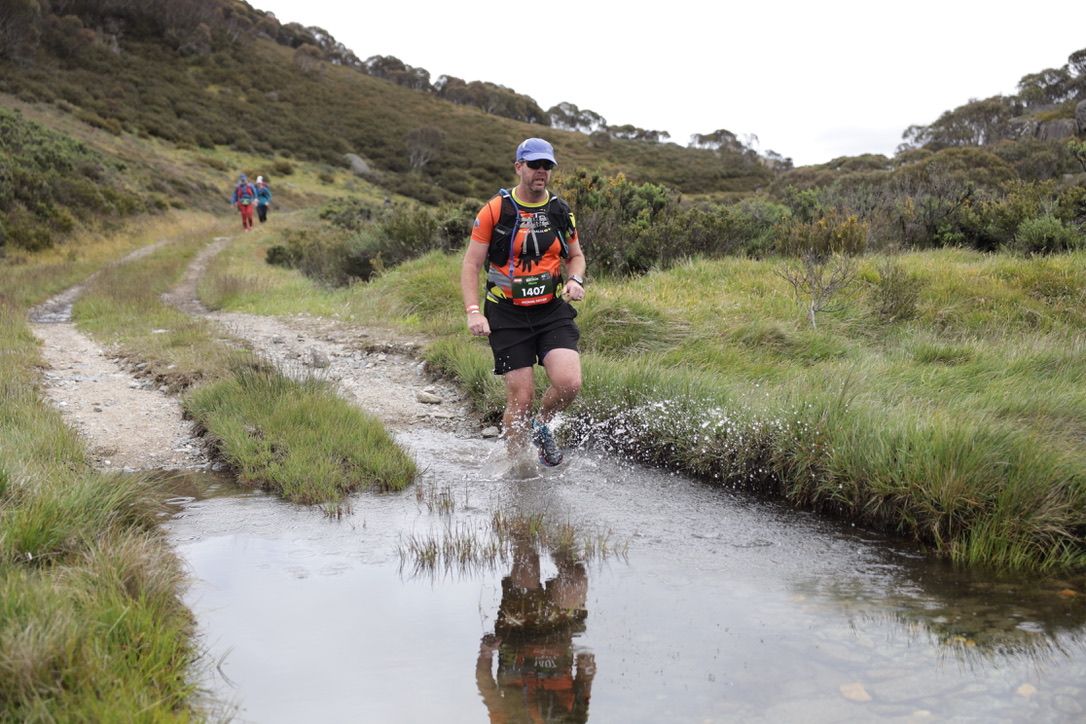 Athlete crossing stream on rocky trail during endurance run.