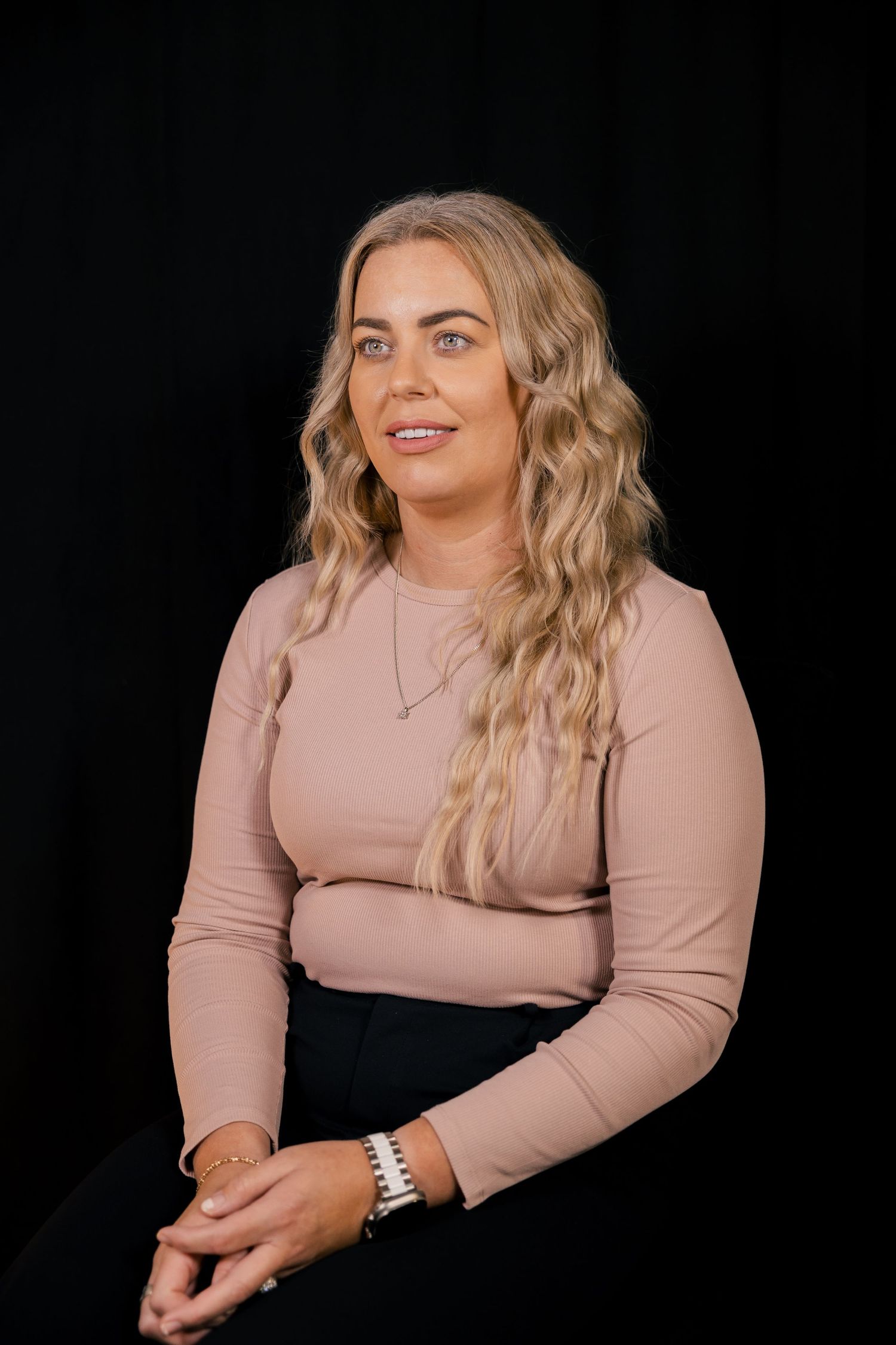 Blonde woman in pink top, seated studio portrait with dark background.