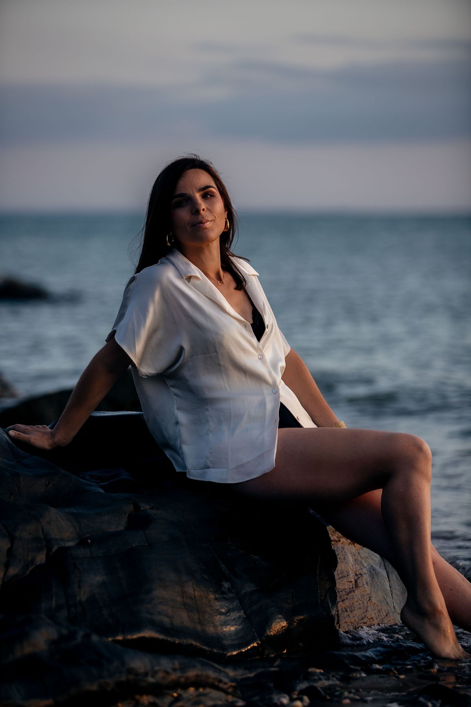 A figure in a white shirt poses on coastal rocks at sunset with the ocean and moody sky in the background.
