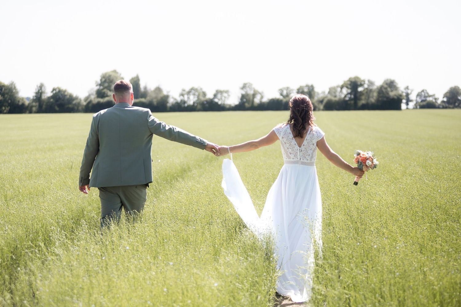 Bride and groom holding hands walking through a lush green field on their wedding day.
