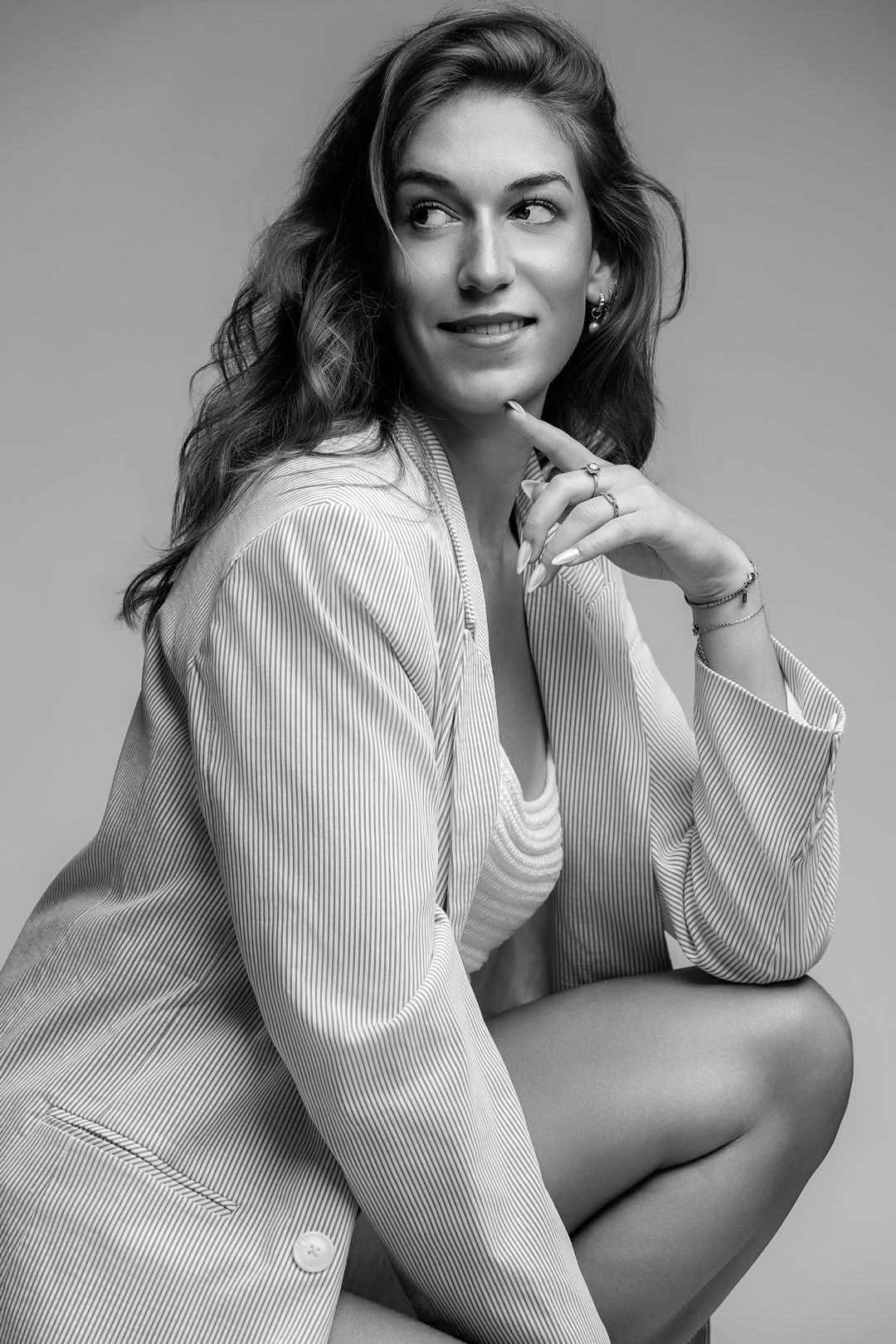 Black and white portrait of a smiling woman in a striped blazer, sitting gracefully with hand on chin.