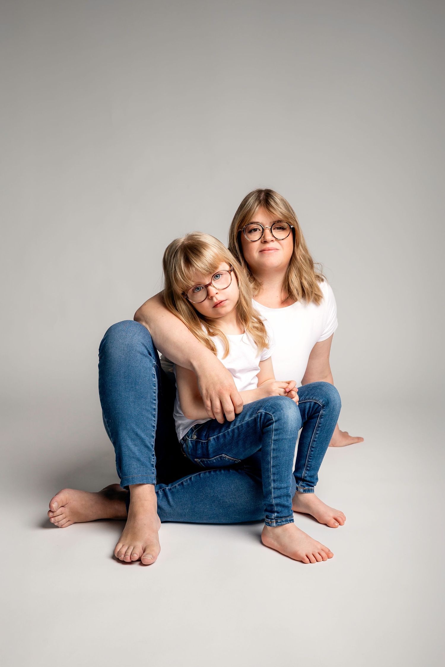 Two young girls in white shirts and jeans sitting together on floor, both wearing glasses, in a studio setting.