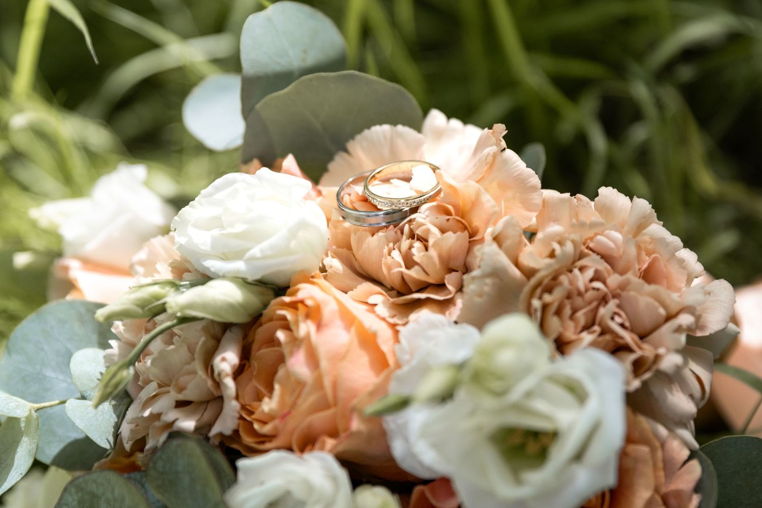 Wedding rings resting on a bridal bouquet of peach roses, white carnations, and eucalyptus leaves.