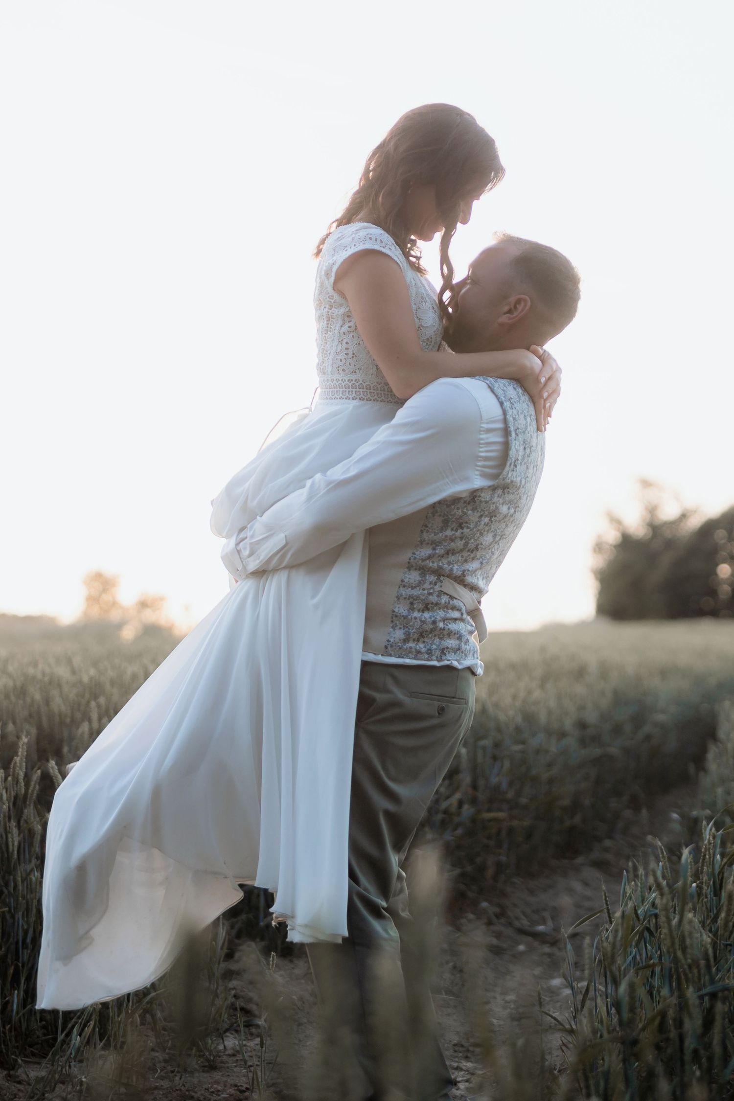 Groom lifting bride in white dress in a golden field at sunset, sharing a romantic moment outdoors.