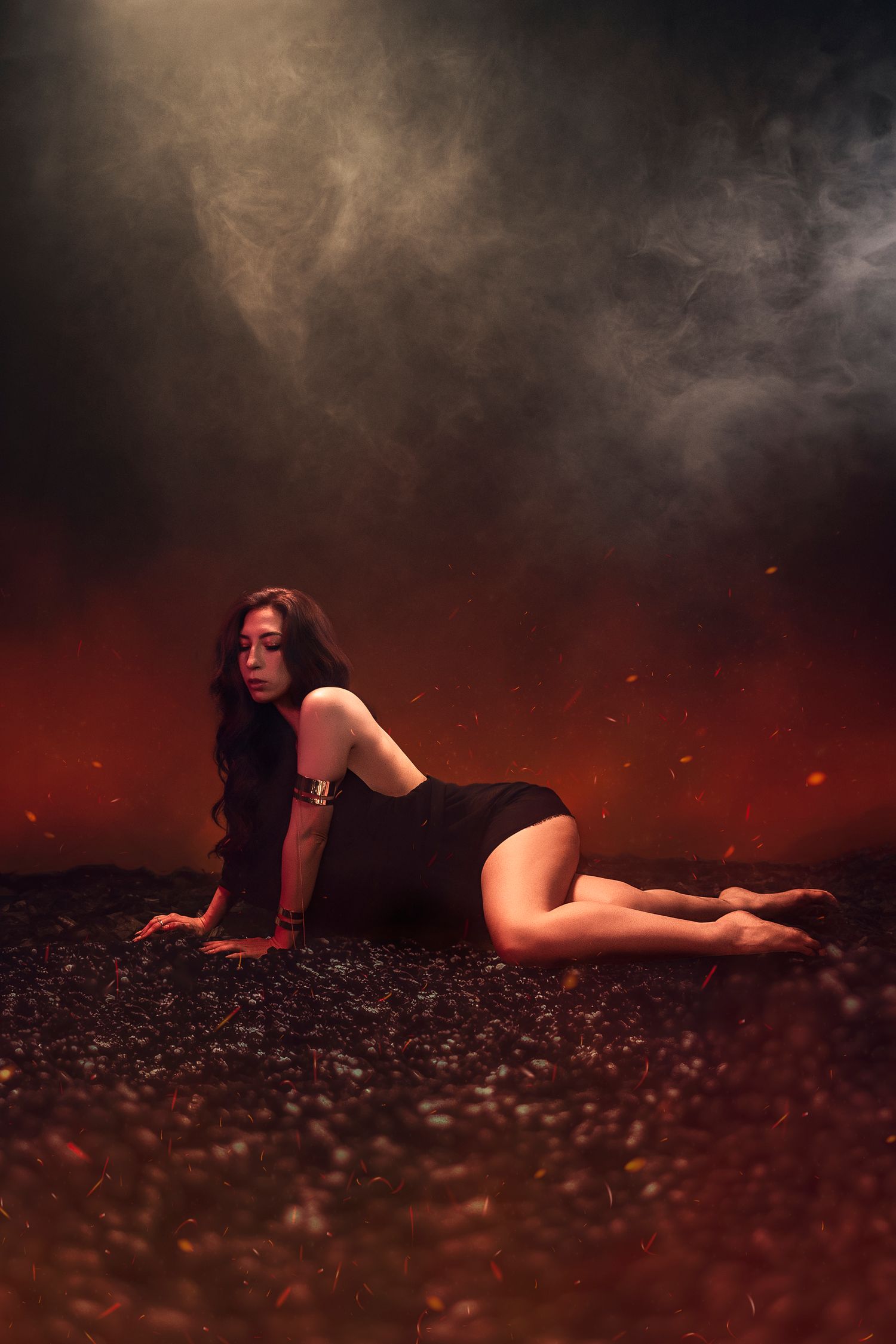 Woman in black dress poses dramatically on dark ground with red flames and smoke in a moody, artistic photo shoot.