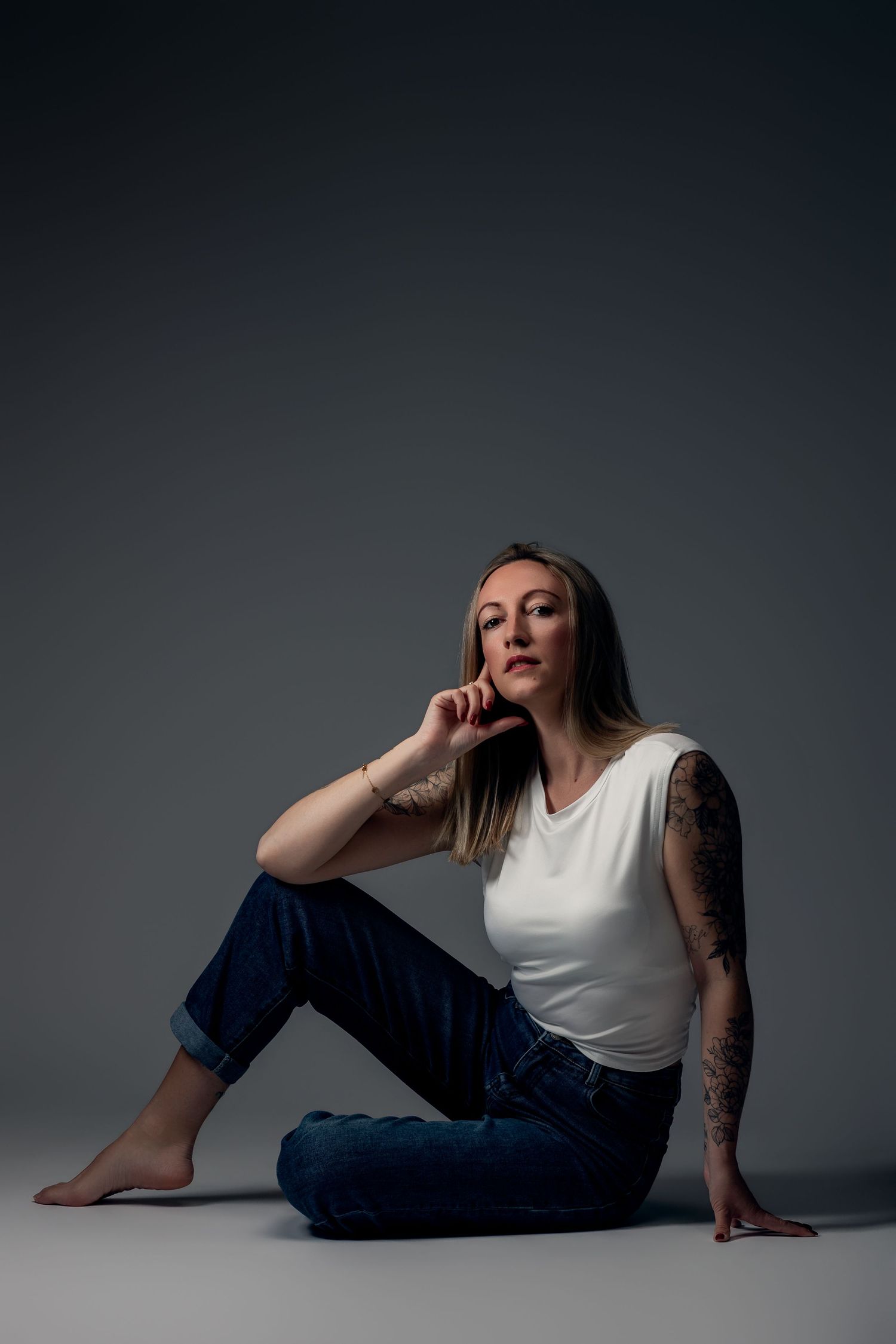 Woman with tattoos sits on floor in white tank top and jeans, posing thoughtfully against gray backdrop.