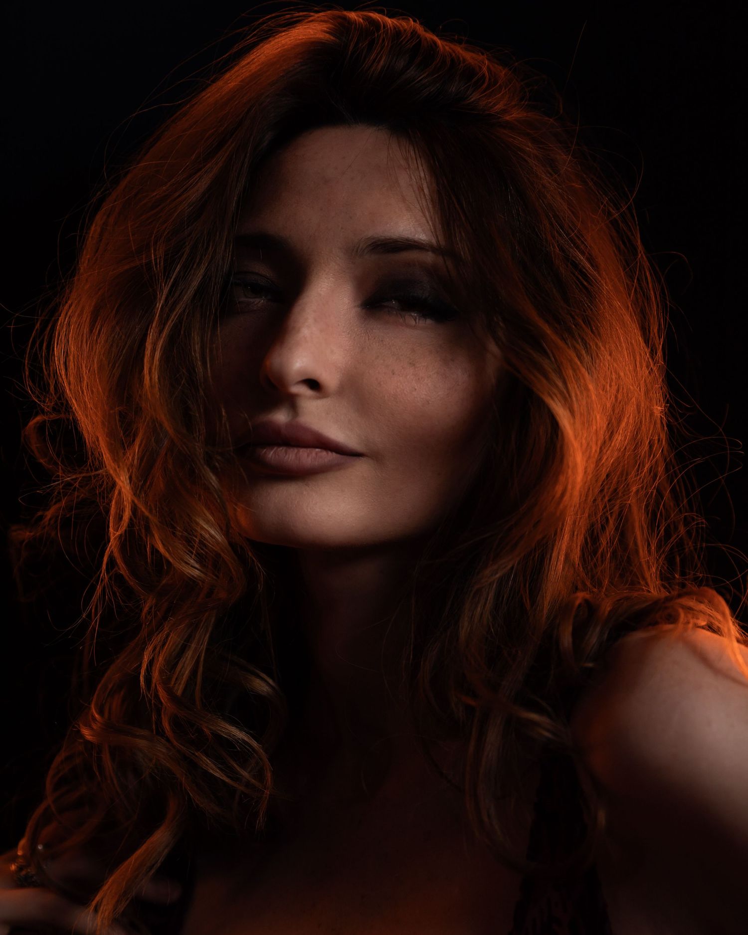 A woman with wavy auburn hair and freckles poses dramatically against a dark background with warm orange lighting.