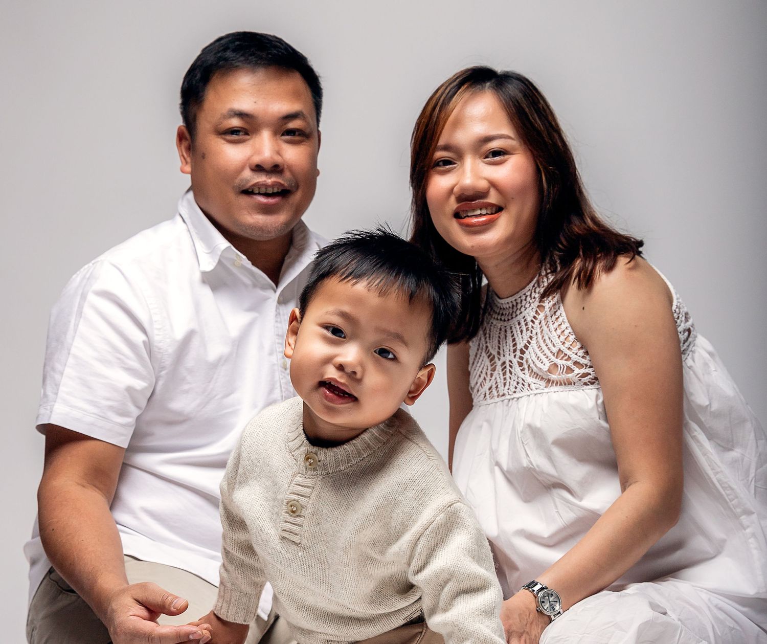 A smiling Asian family with a father, mother, and young toddler son posing together in a bright studio.