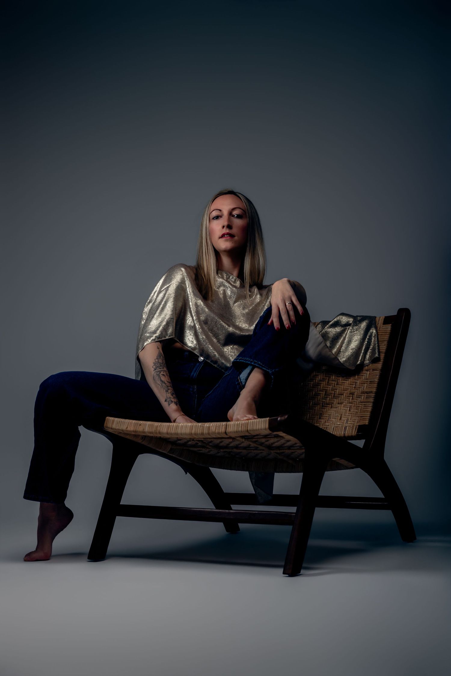 Woman in gold top and navy pants sits barefoot on wicker chair, showcasing tattoo sleeve in moody studio lighting.