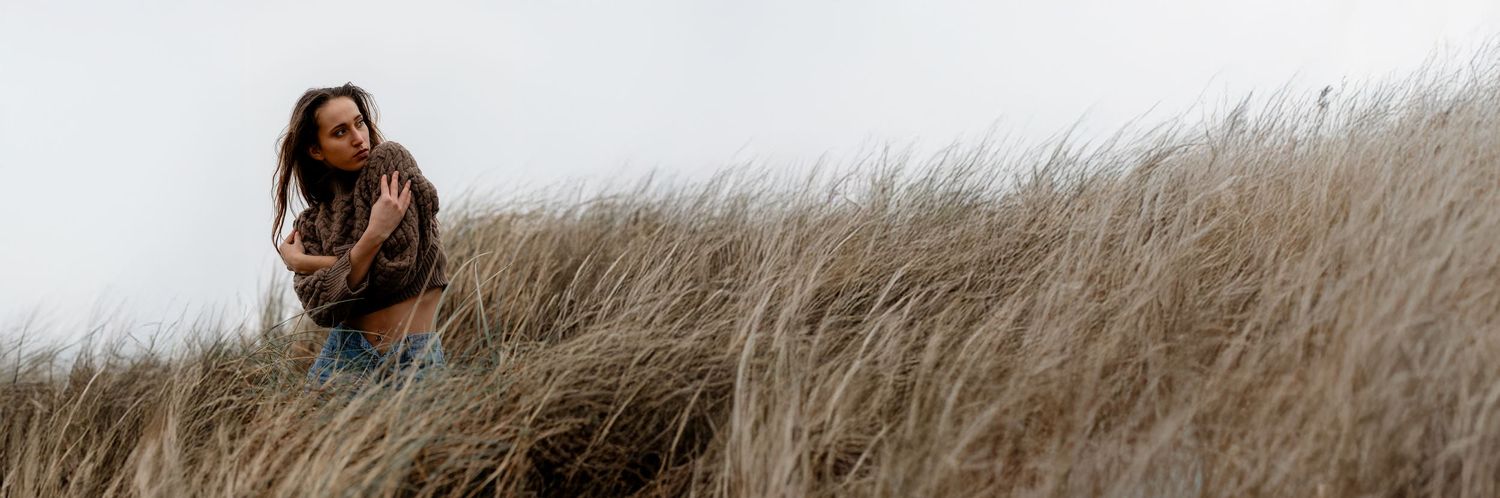 A lone silhouette stands among tall windswept grasses on a hilltop against an overcast sky.