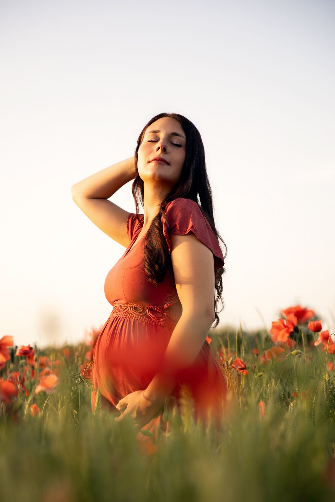 Pregnant woman in red dress standing in a blooming poppy field at golden hour with eyes closed.