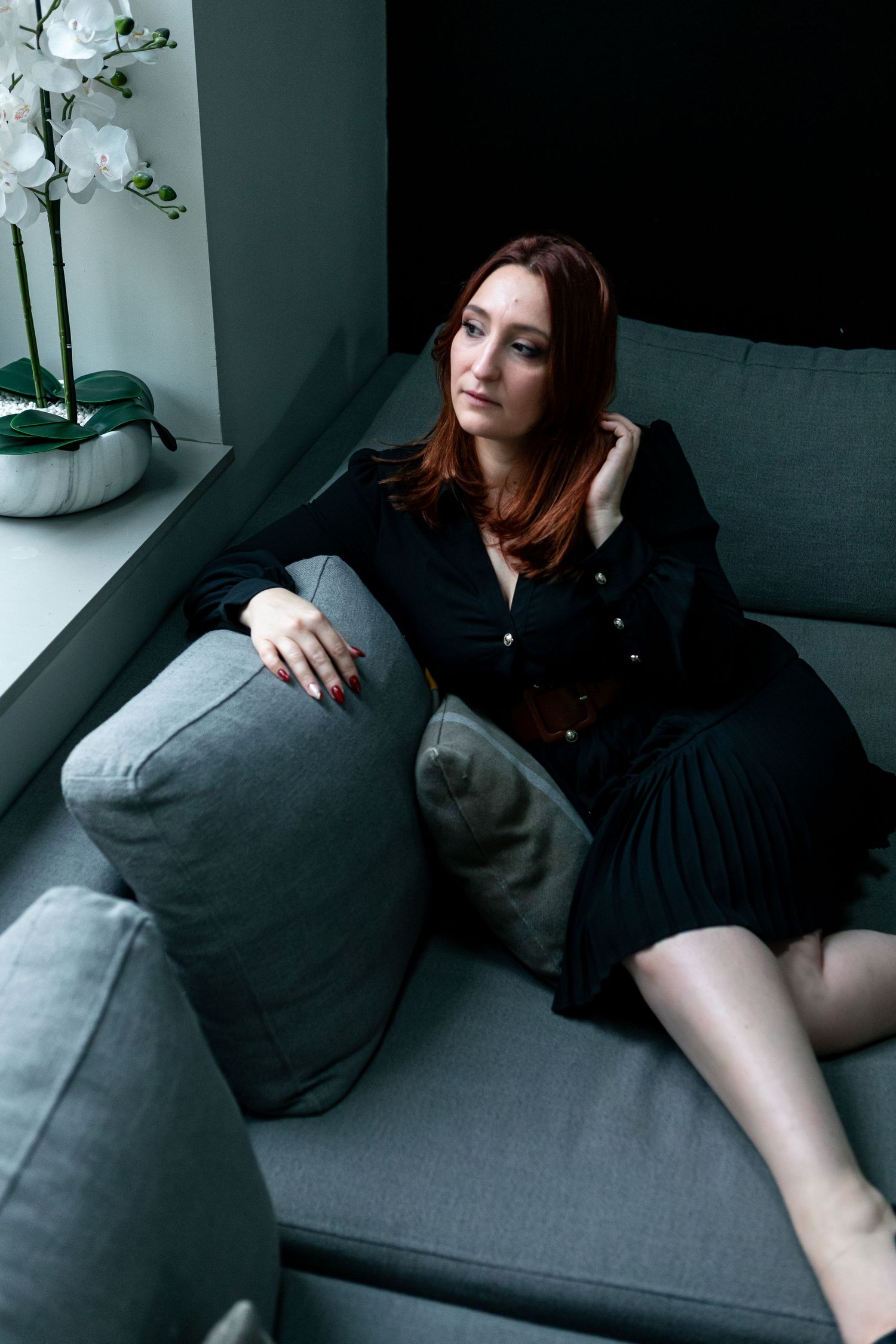 A woman in dark clothing relaxes on a gray couch near a window with white orchids.