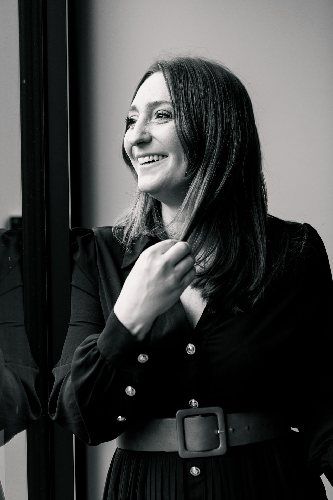 Black and white portrait of a smiling woman in a belted black dress with silver buttons, posing by a window.