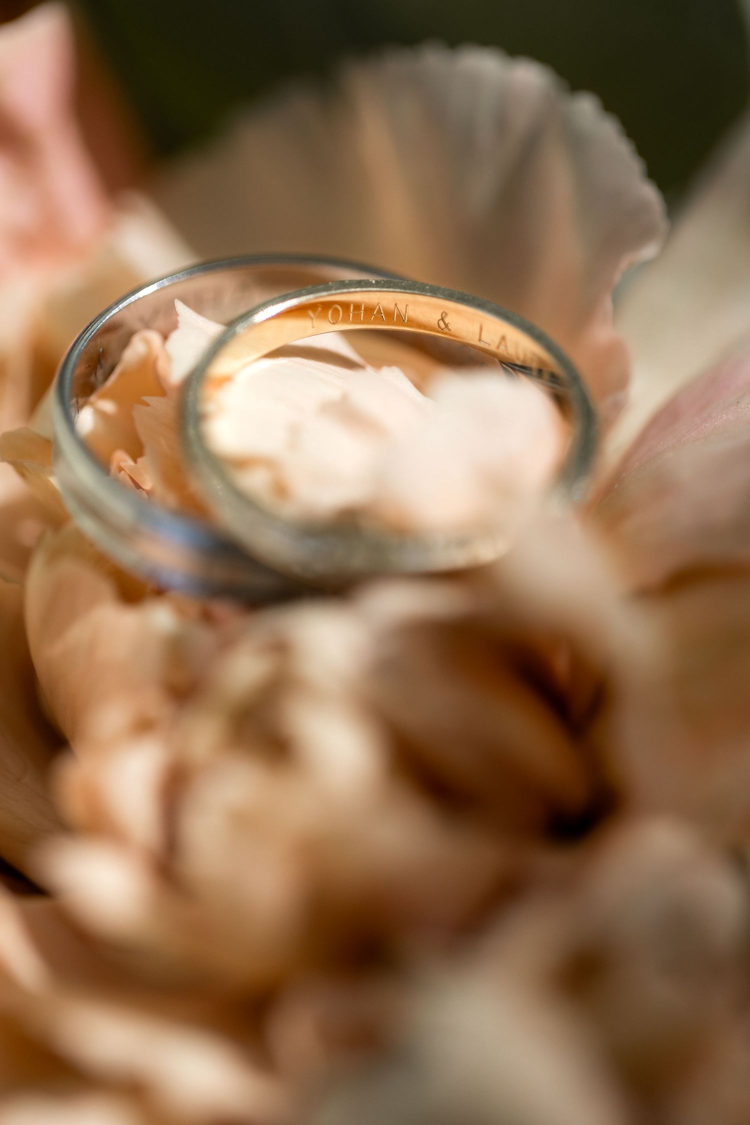 Two gold wedding rings with engraved names rest inside a glass jar nestled among soft pink peony petals.