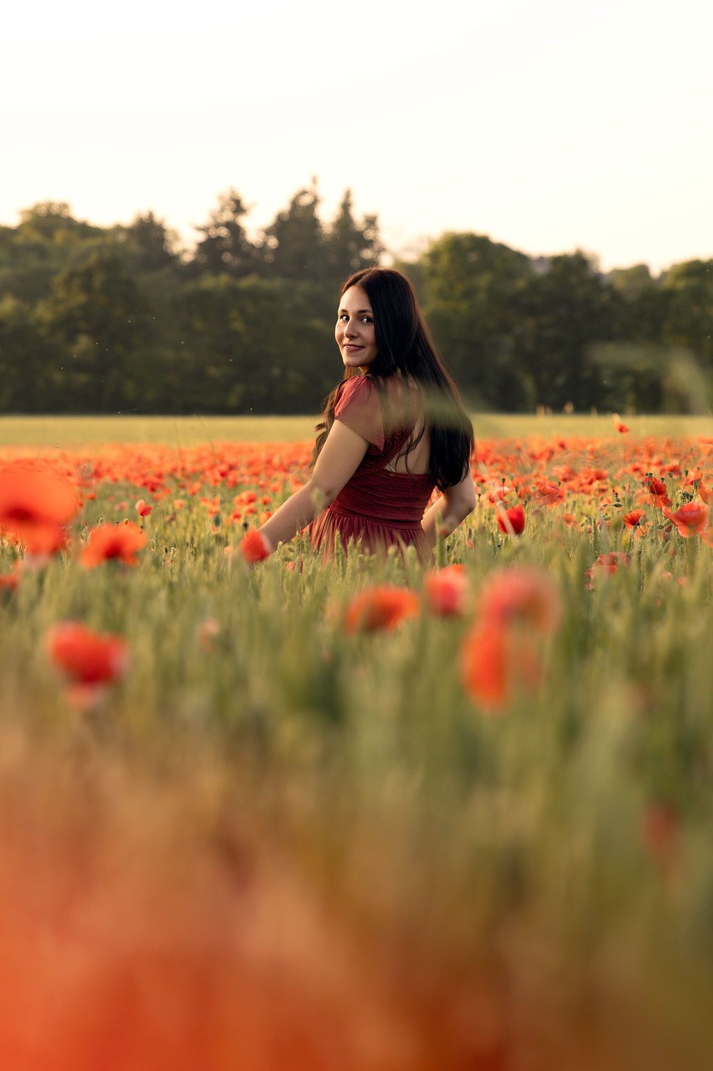 A smiling brunette woman in a red dress standing in a vibrant poppy field at golden hour.