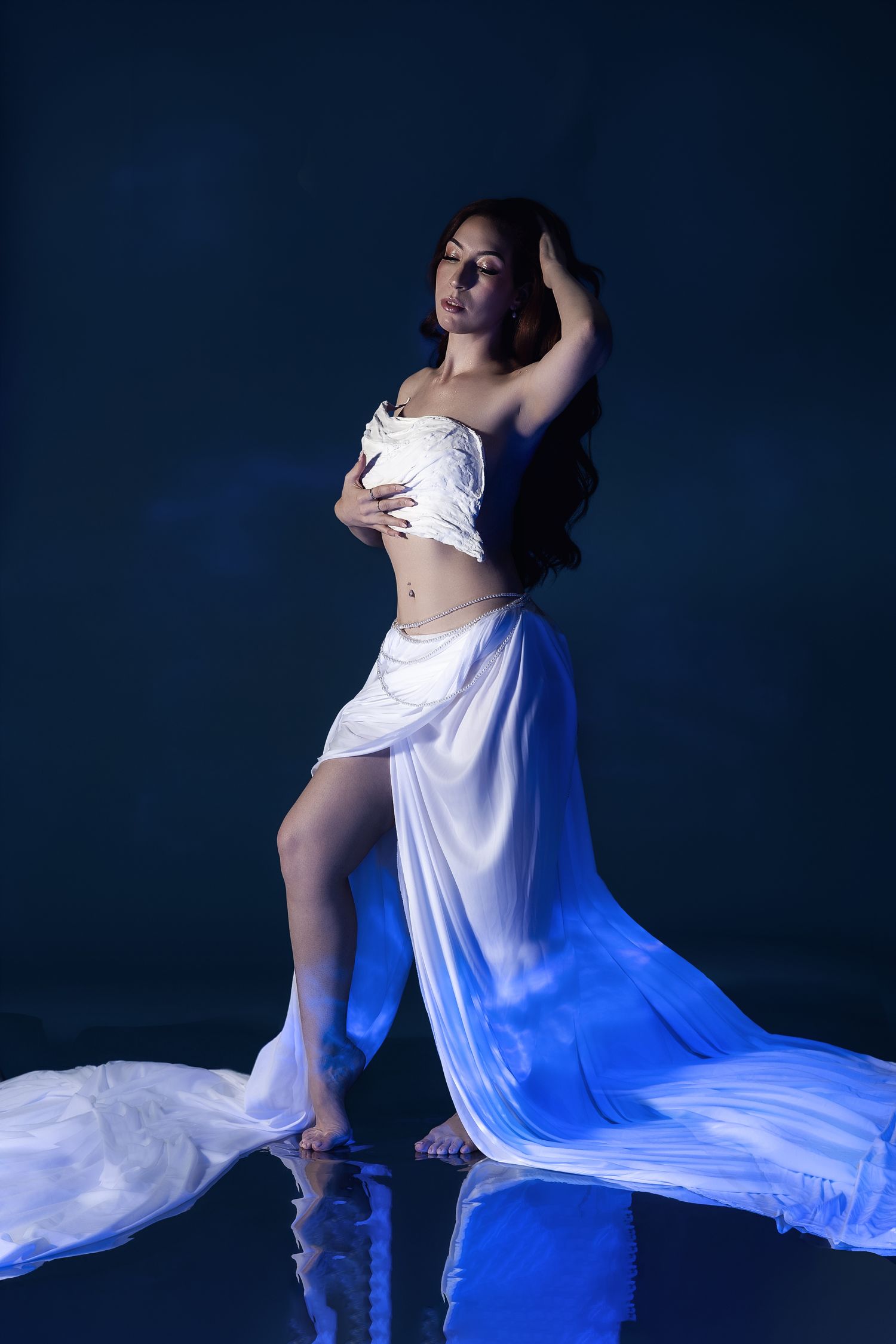 A woman in a flowing white dress poses gracefully against a dark blue background with dramatic lighting.