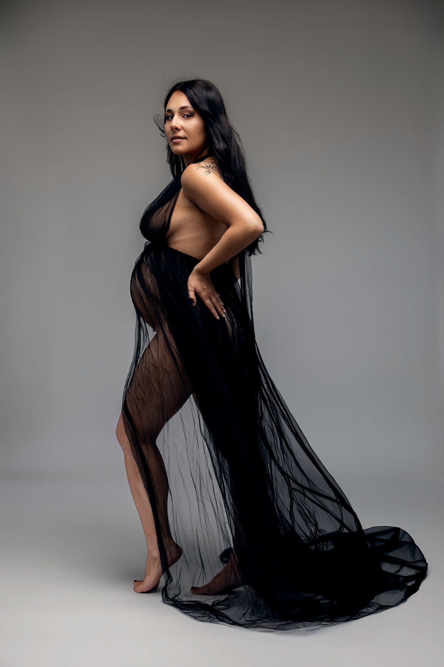 Pregnant woman in sheer black flowing gown posing gracefully against gray studio background.
